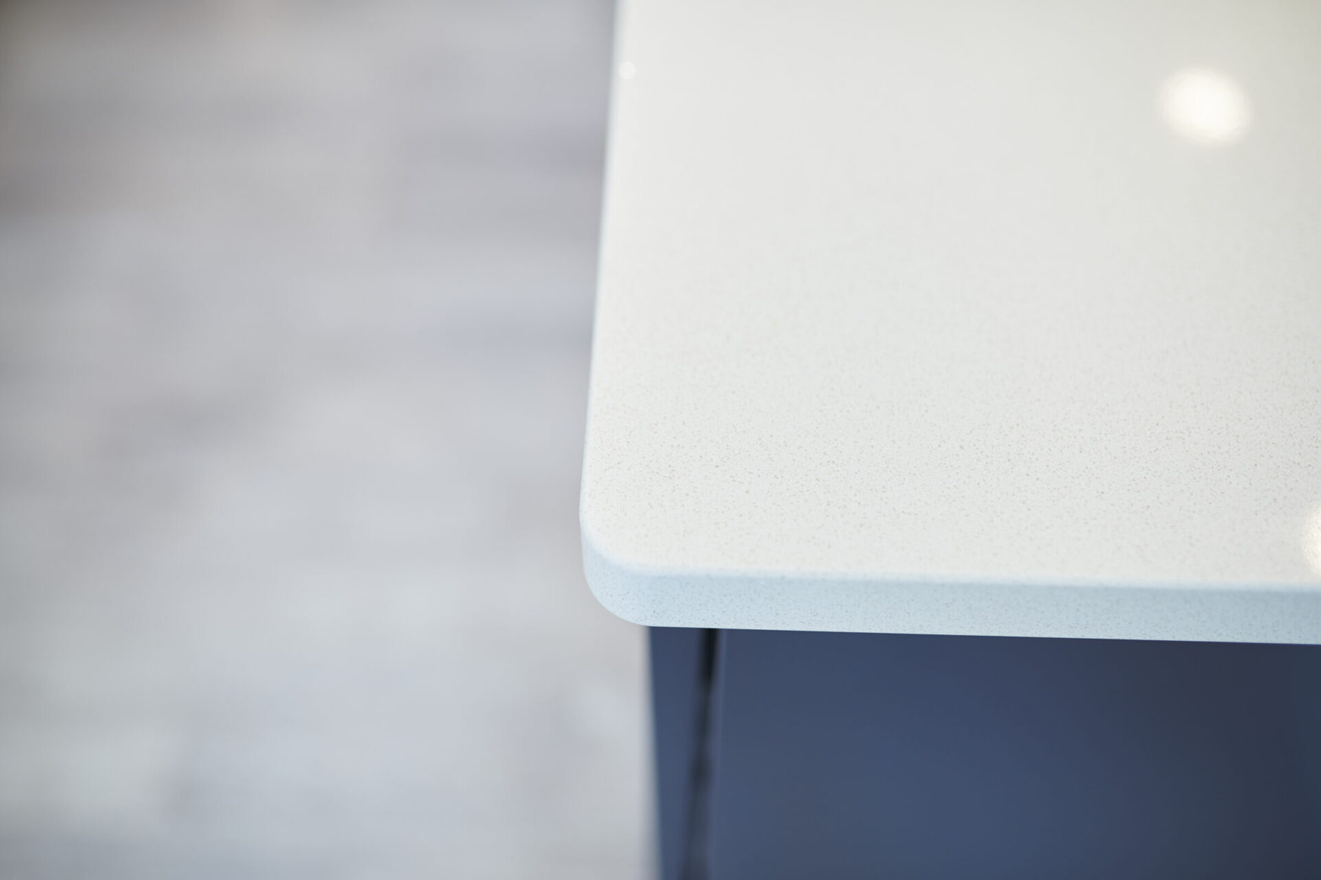 This is a close-up image of a modern, minimalist table with a speckled white tabletop and a blurred background, focusing on the corner edge.