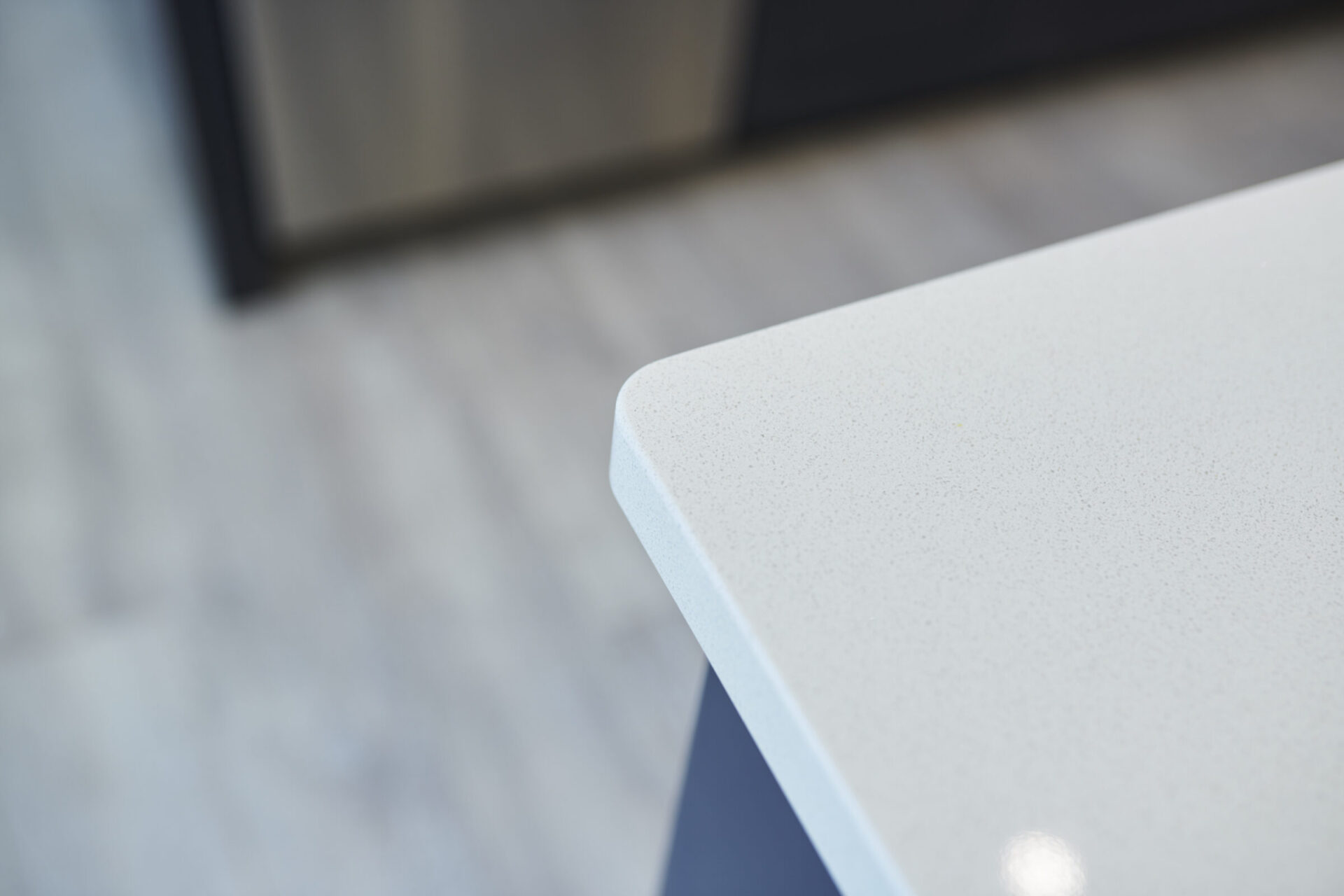 This image showcases a close-up of a modern kitchen counter's corner with a speckled white surface, against a blurred background of cabinetry and flooring.