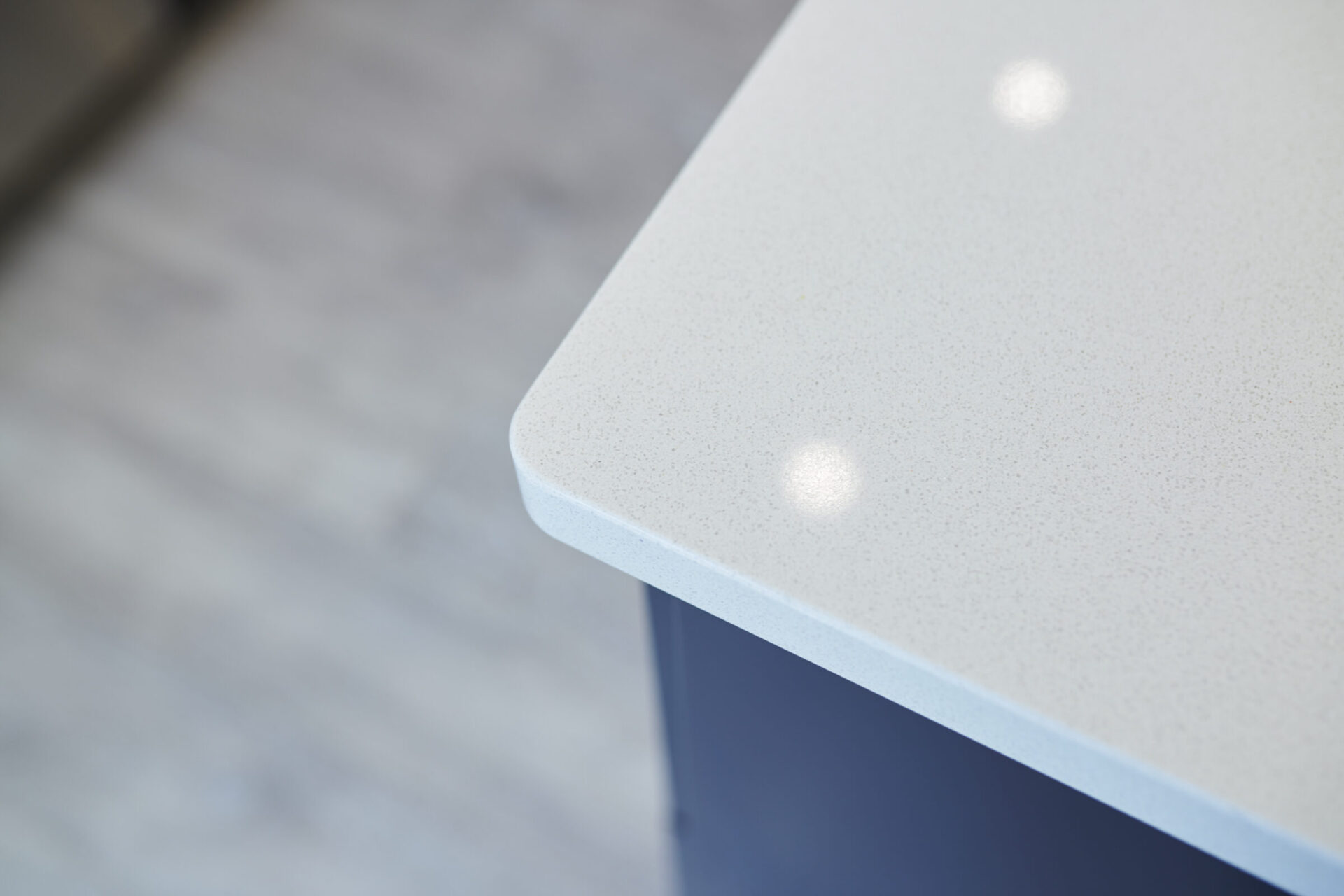 The image shows a close-up corner of a minimalist kitchen countertop with a speckled surface, reflecting overhead lights, atop a navy cabinet base.