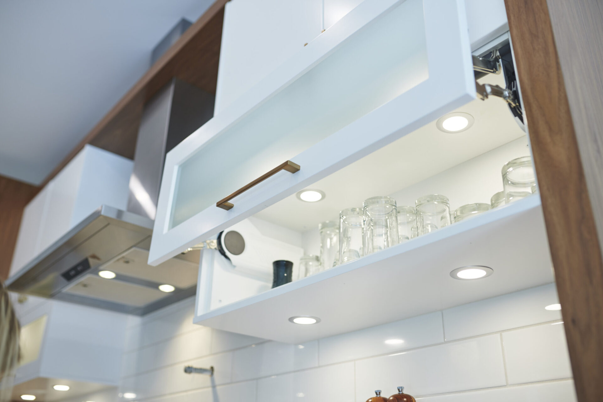 An open upper kitchen cabinet with clear glass jars. The white cabinet has modern handles and built-in lights, against a tiled backsplash.