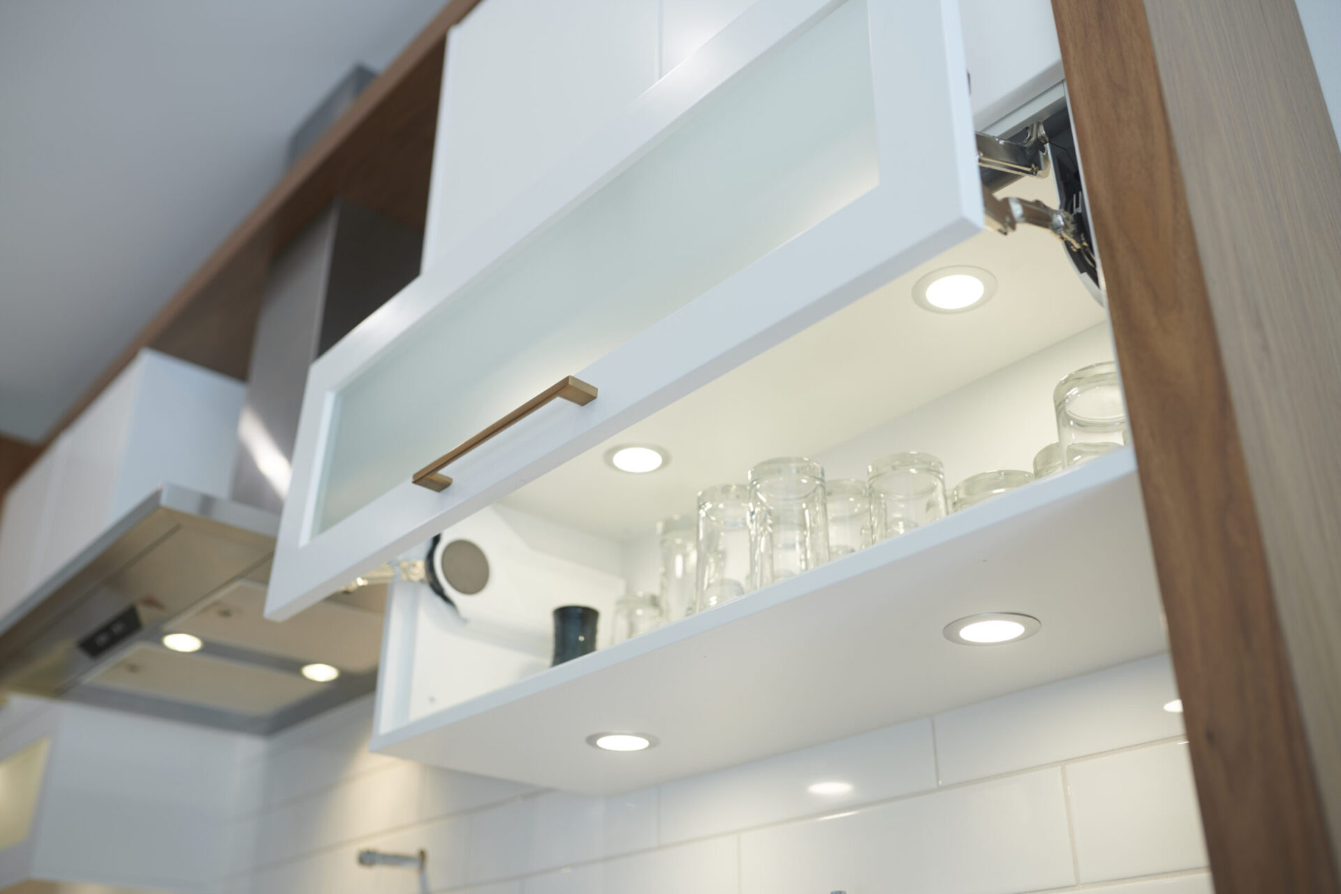 Modern kitchen cabinet with open frosted-glass door revealing neatly organized clear glasses. Bright interior lighting highlights clean, white tiles and wooden accents.