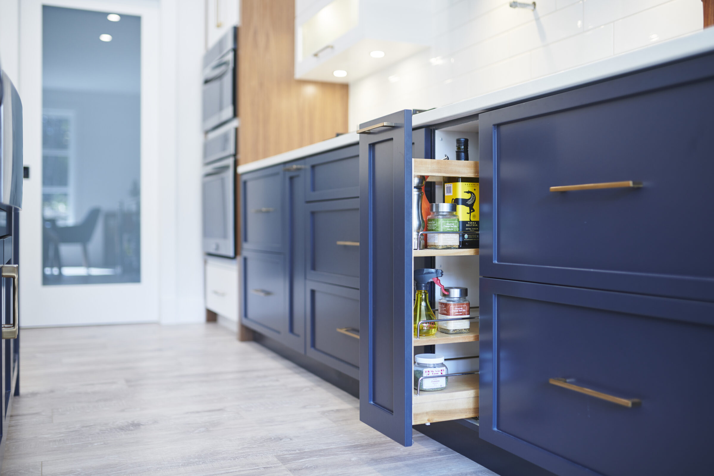 Modern kitchen interior with blue cabinets, gold handles, and a pull-out organizer displaying spices and oils. Light wood flooring complements the clean design.