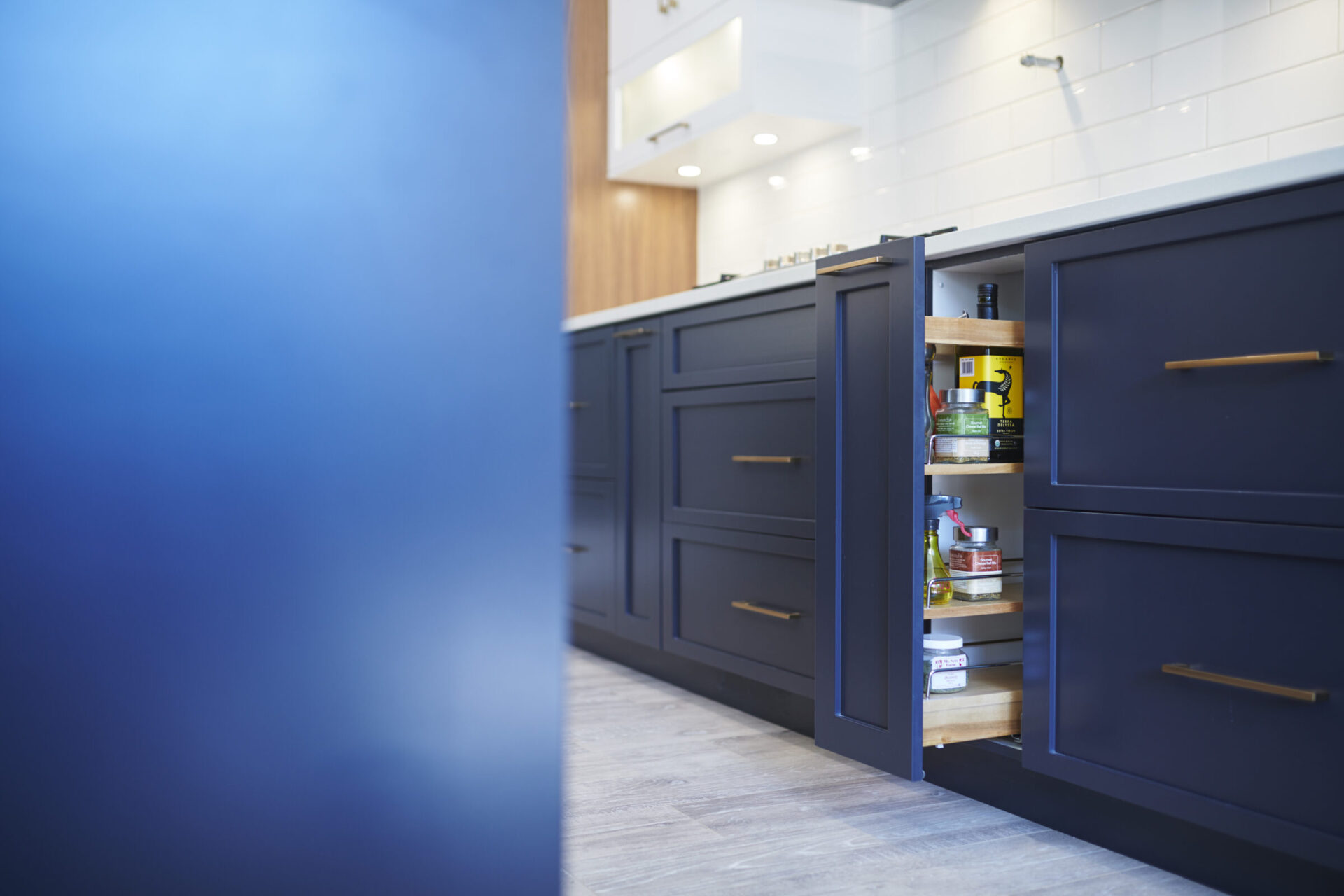 Modern kitchen with navy blue cabinets, brass handles, and an open drawer revealing organized spices and condiments. Light wooden elements and white tiles accent the space.