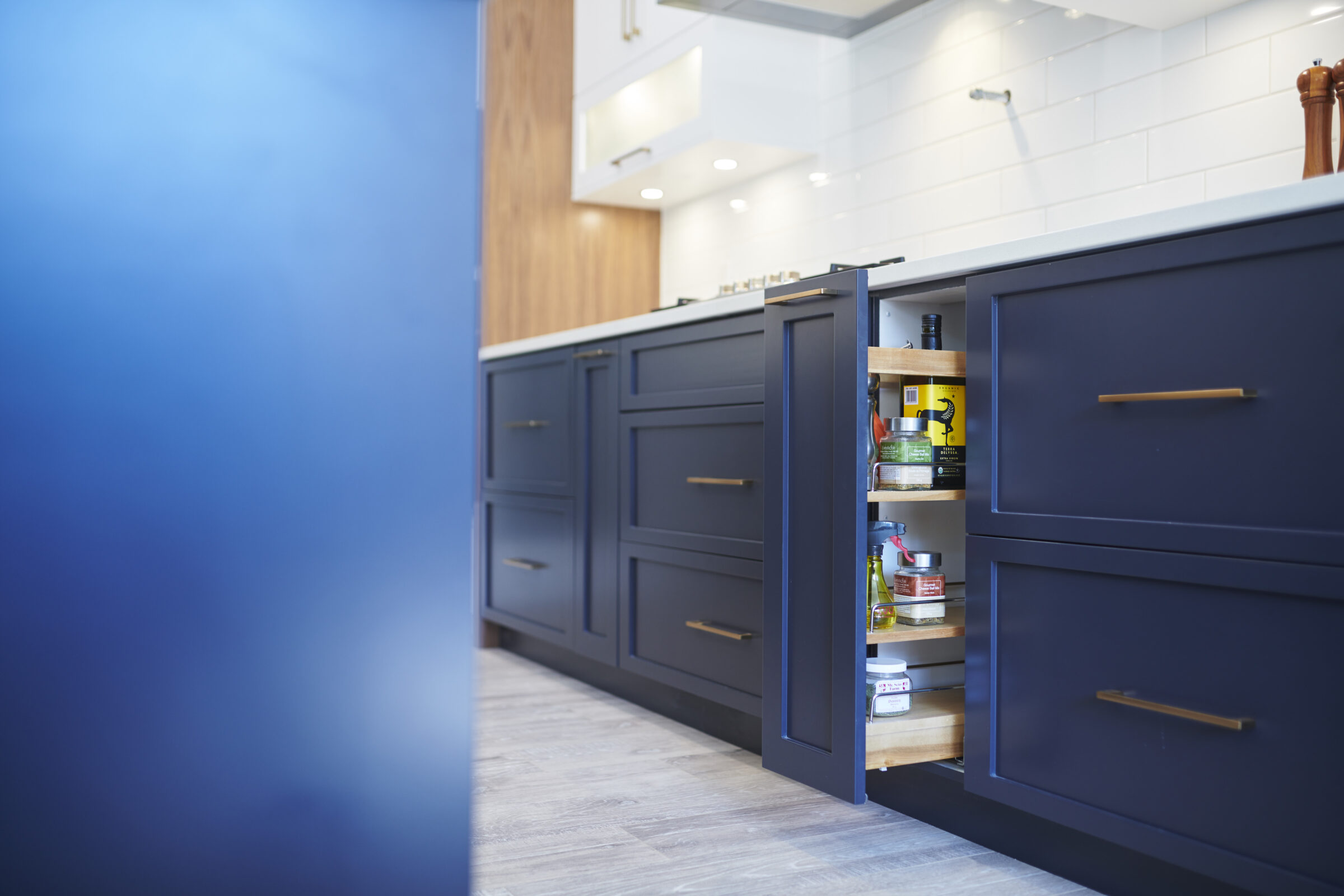 Modern kitchen interior with navy blue cabinets, brass handles, and wooden countertops. Open cupboard reveals organized spices and condiments. Bright, clean atmosphere.