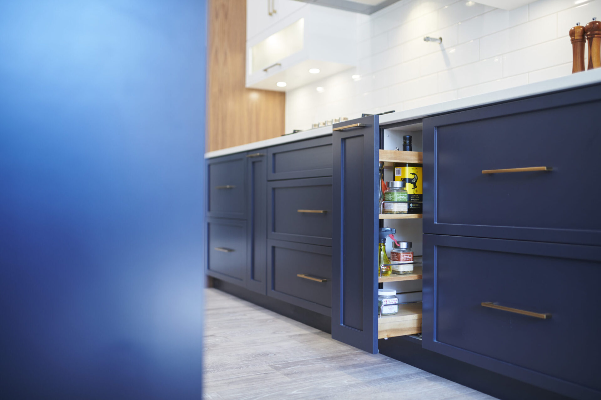 A modern kitchen with dark blue cabinets, one open revealing organized shelves filled with condiments, under warm lighting against a subway tile backsplash.