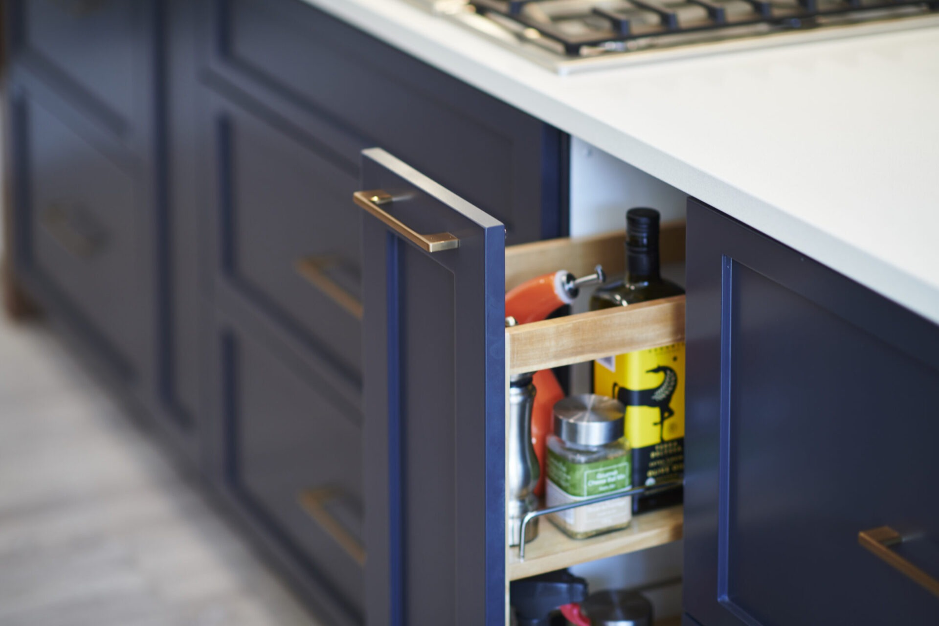 A modern kitchen with navy blue cabinets featuring gold handles, a white countertop, and a pull-out storage rack with various condiments and bottles.