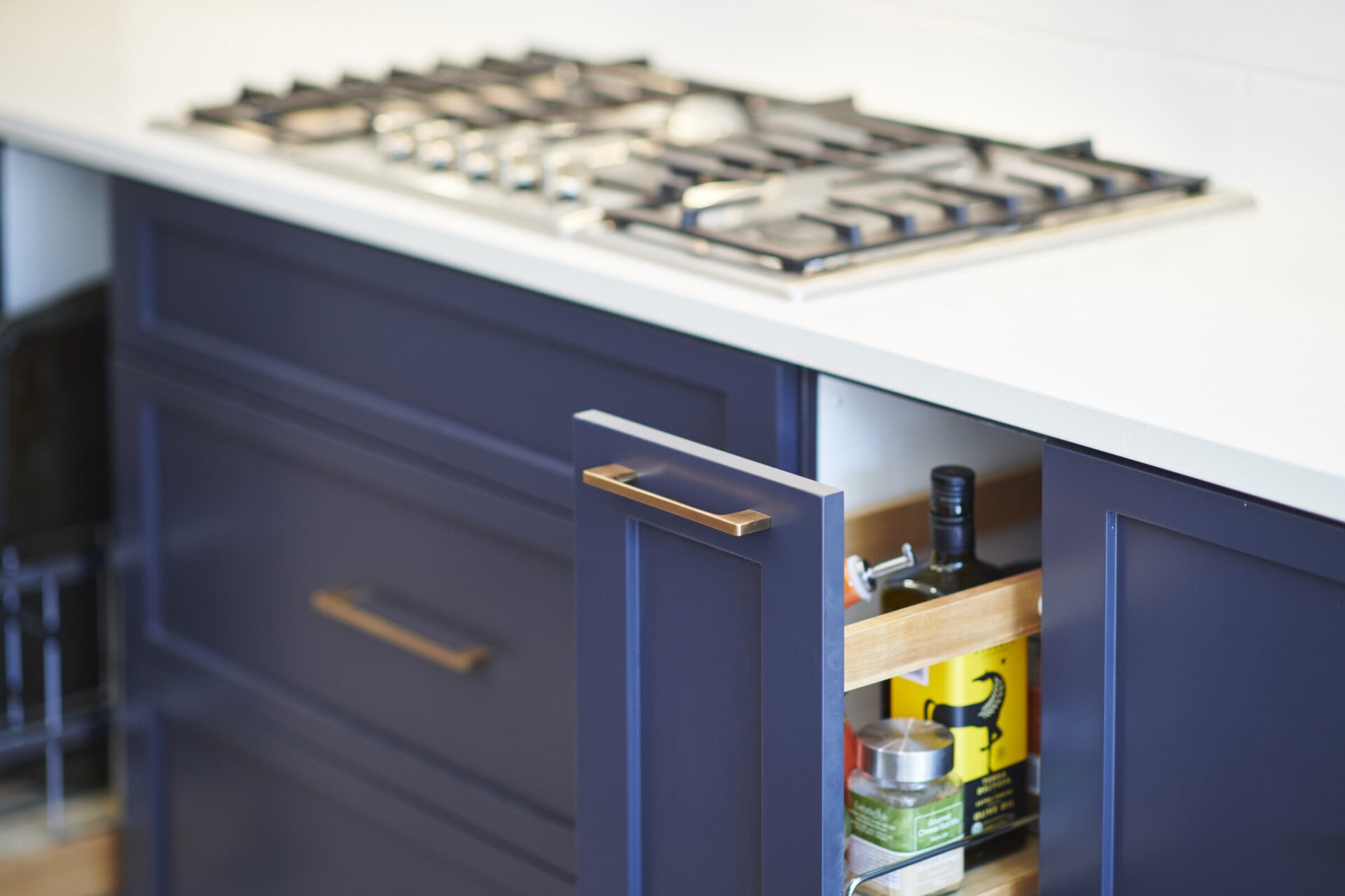 A modern kitchen with a gas stovetop on a white countertop above navy blue cabinets, one open revealing condiments and a pull-out rack.