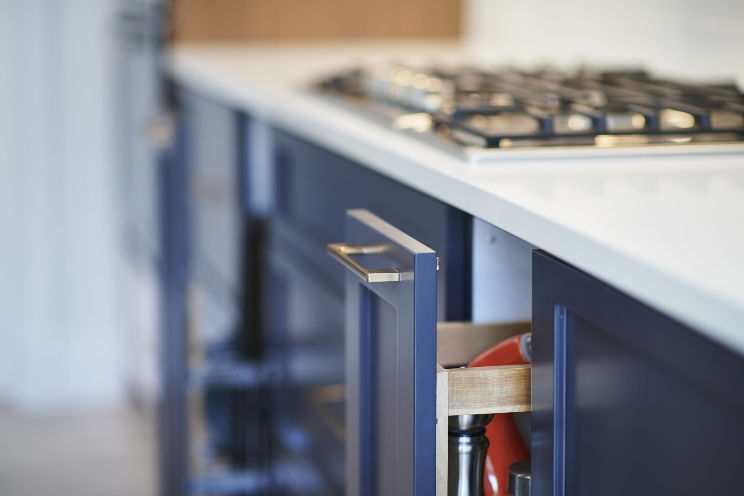 Modern kitchen interior focused on a drawer partially open, revealing cooking utensils. A gas stove top is visible in the blurred background.