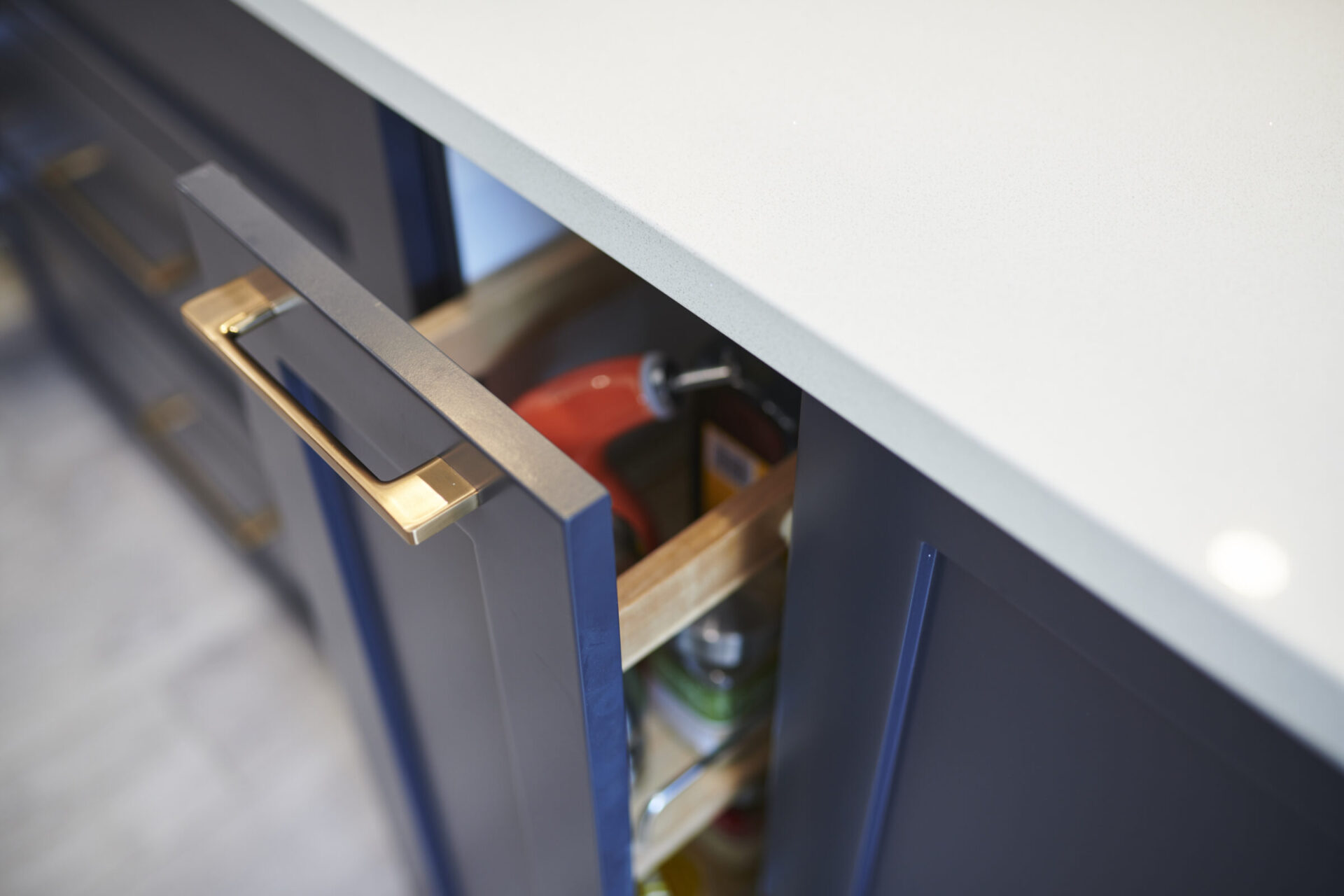 A partially open drawer in a blue kitchen cabinet with a silver handle showing bottles and containers inside. The countertop is light grey.