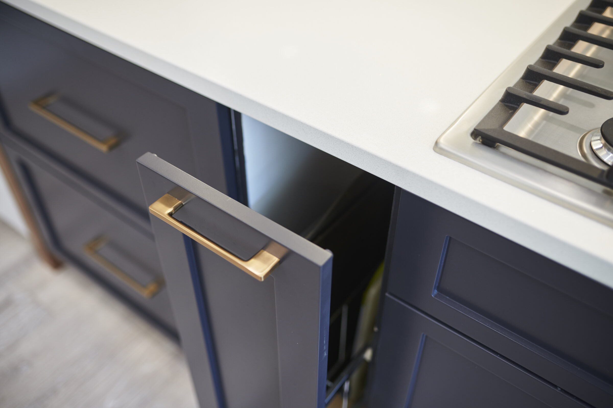 A modern kitchen with a white countertop, navy blue cabinets, and a stainless steel gas cooktop. An open drawer reveals a nested cutlery organizer.