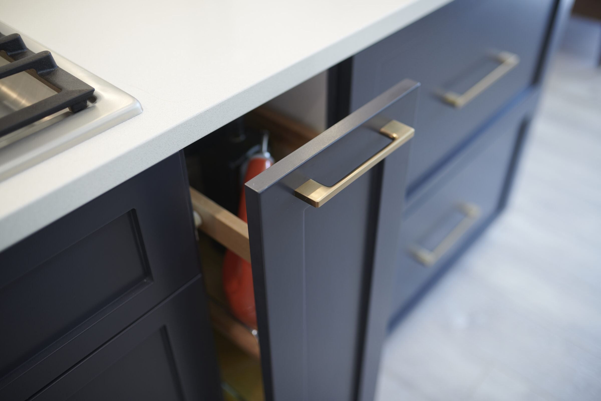 A modern kitchen cabinet with a brass handle is open, revealing contents like a red pot inside. There's a white countertop and gas stovetop visible.