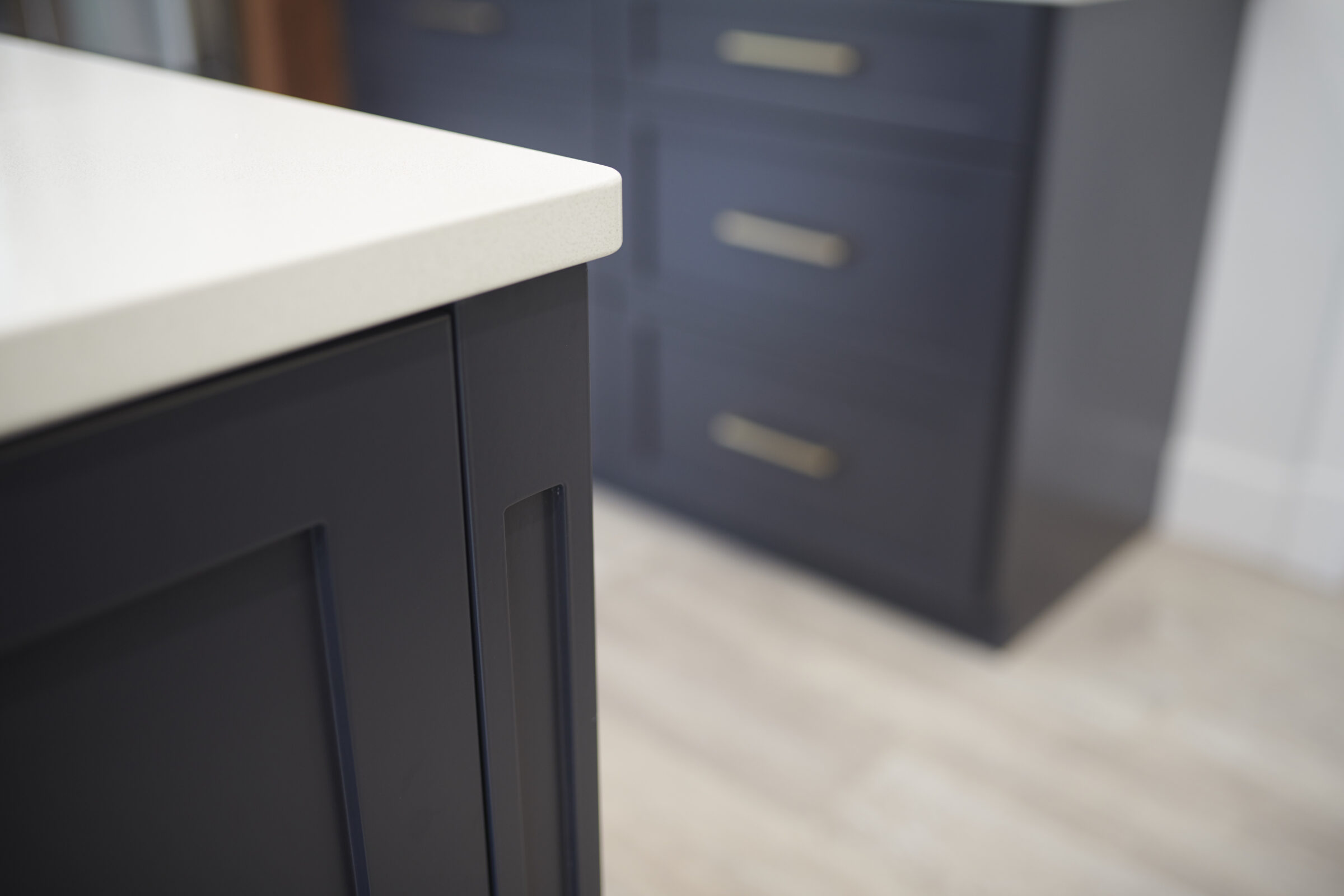 This image shows a close-up of a modern kitchen counter edge with a blurred background featuring dark cabinetry and sleek hardware against a light floor.