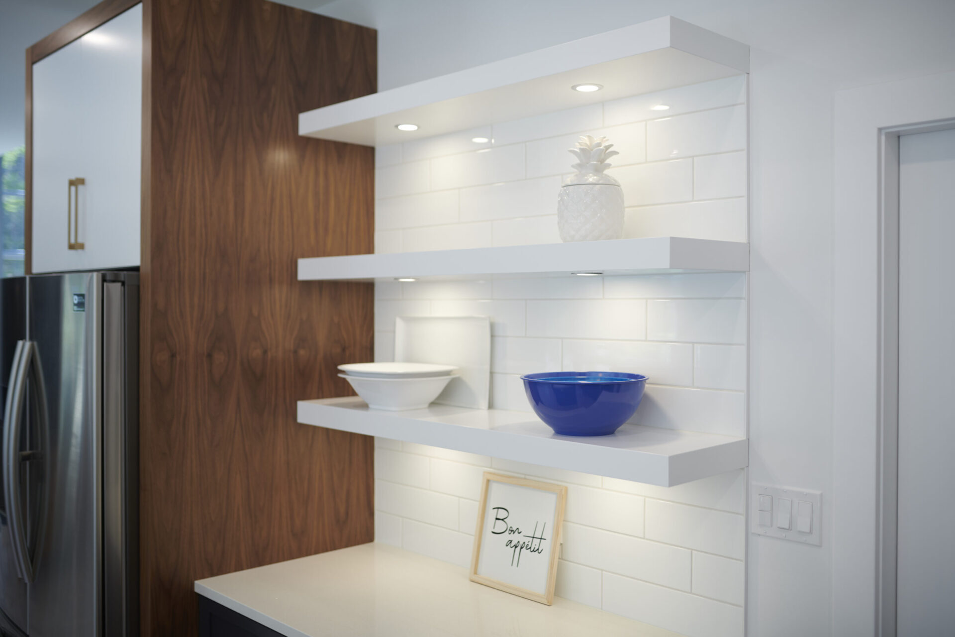 A modern kitchen corner with white subway tiles, floating shelves displaying a pineapple decoration, blue bowls, and a 