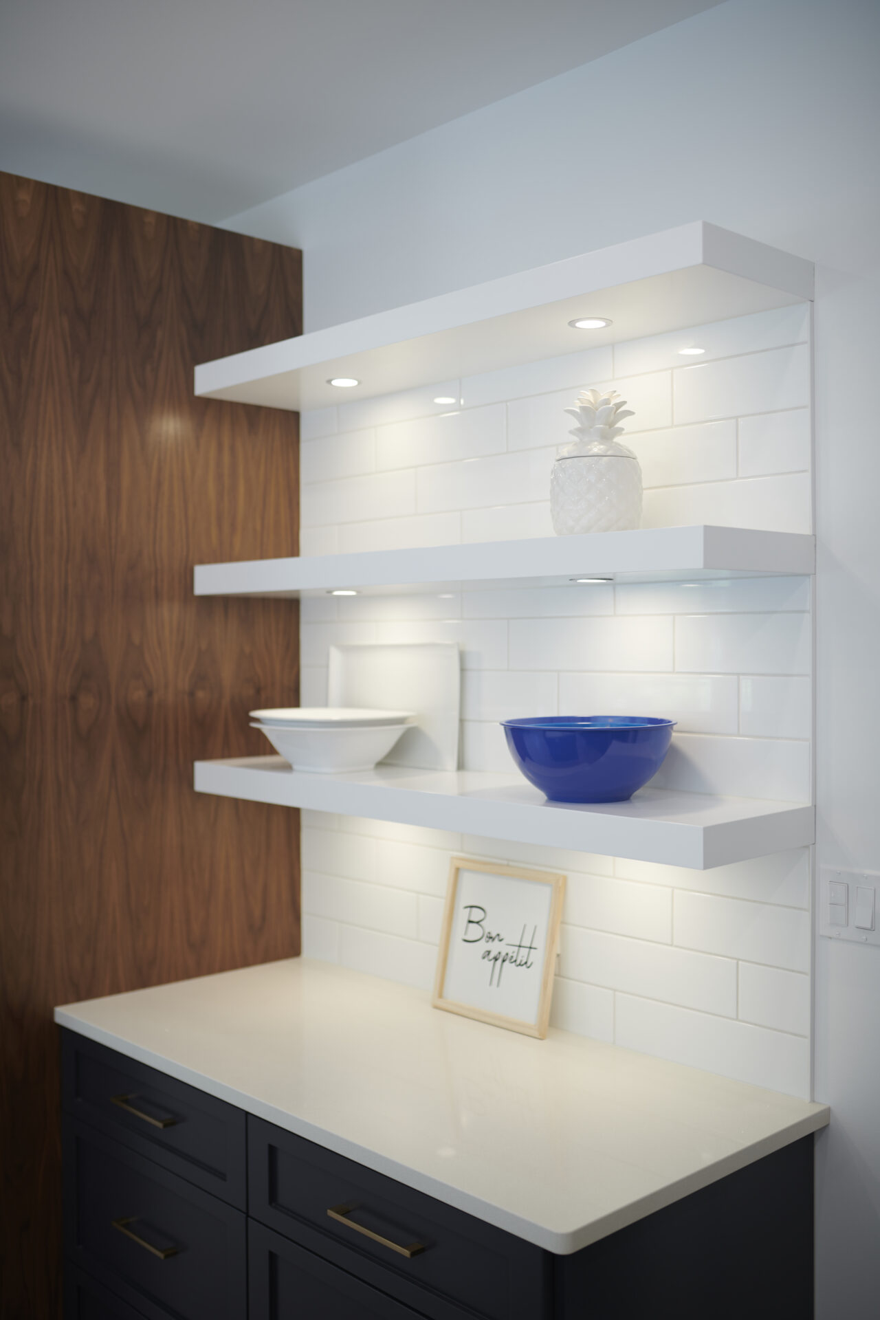 This image shows a modern kitchen corner with white floating shelves, a wooden wall, decorative items, and a dark blue cabinet with a countertop.