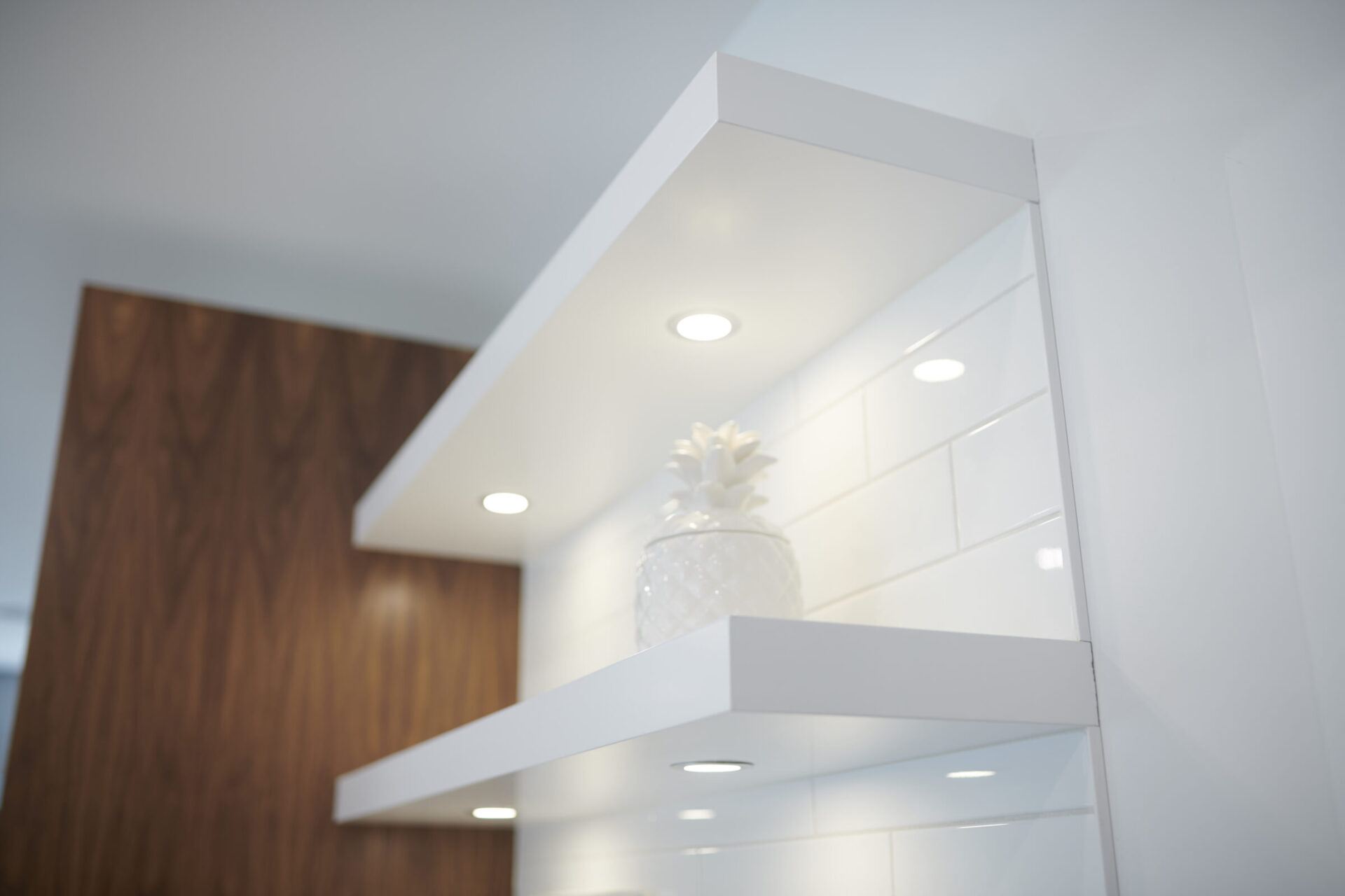 This image features a modern interior design with sleek white shelves, recessed lighting, and a decorative pineapple accent against a contrasting wood panel wall.