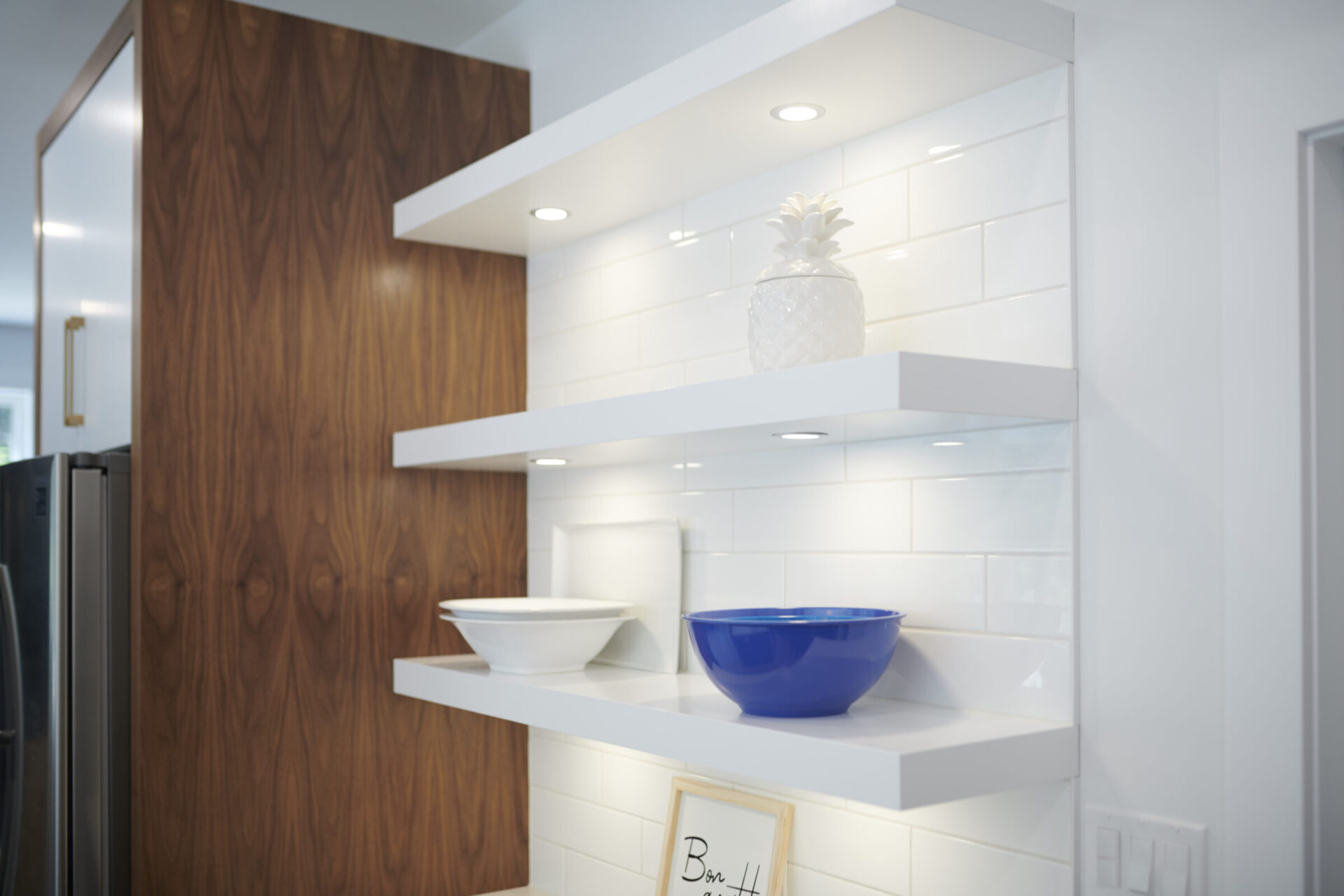 This image shows a modern kitchen corner with white open shelves. A blue bowl, white dishes, and a decorative pineapple adorn the shelves against a wood panel.