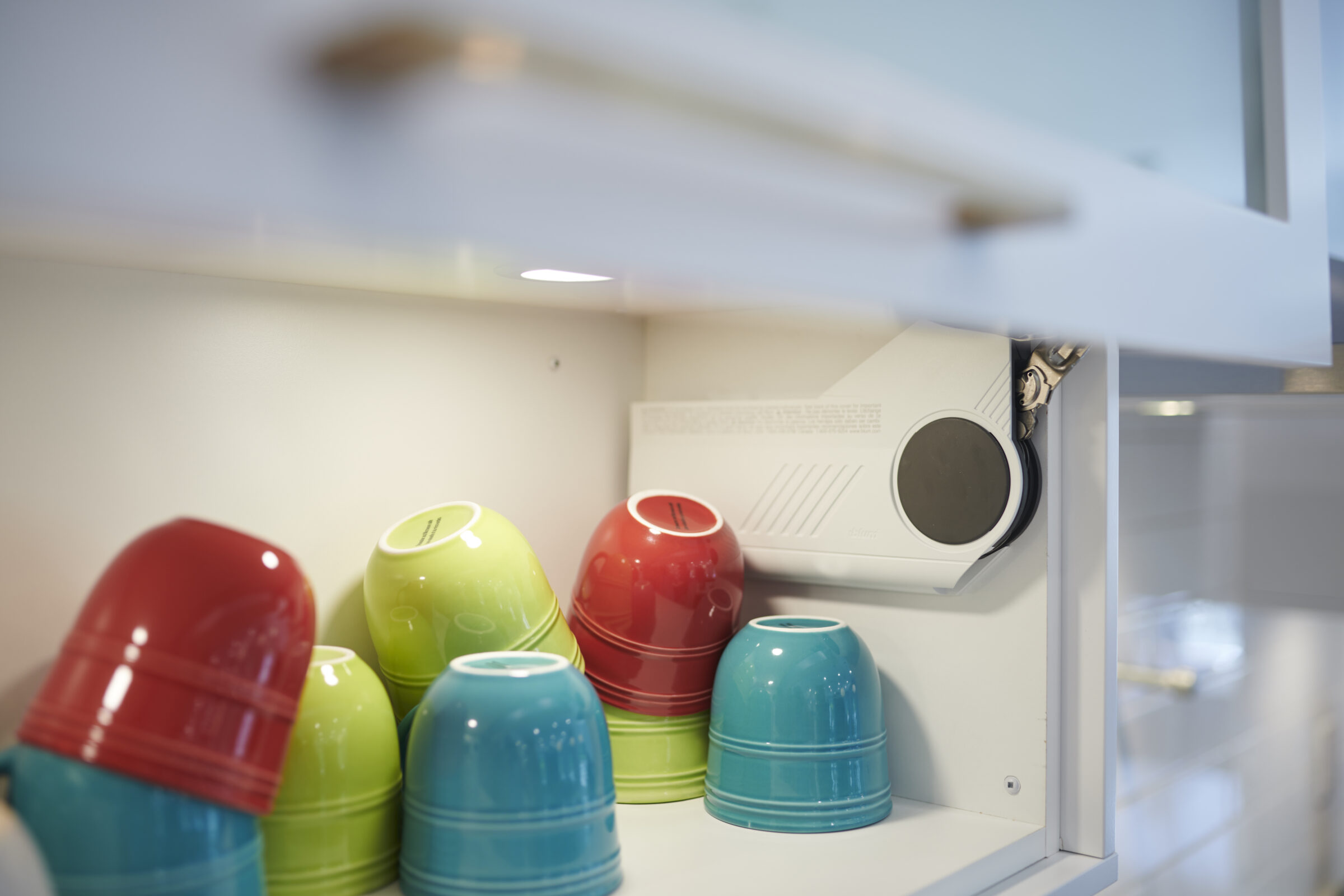 The image shows a modern kitchen shelf with colorful upside-down bowls next to a white built-in projector, suggesting a space-conscious, tech-integrated home environment.