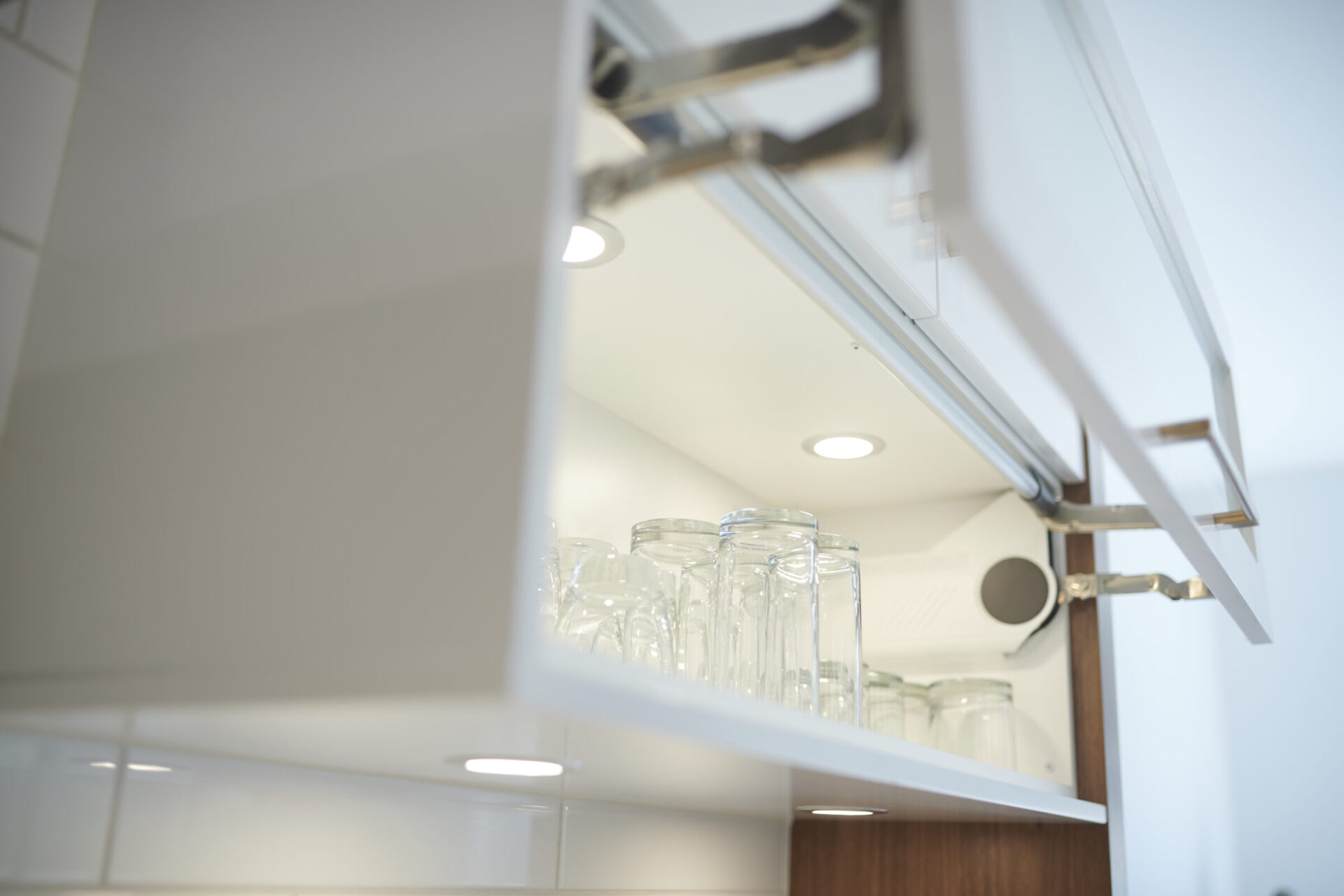 This image features an open white cabinet with clear glass tumblers neatly arranged on a shelf, illuminated by recessed lighting in a clean kitchen.