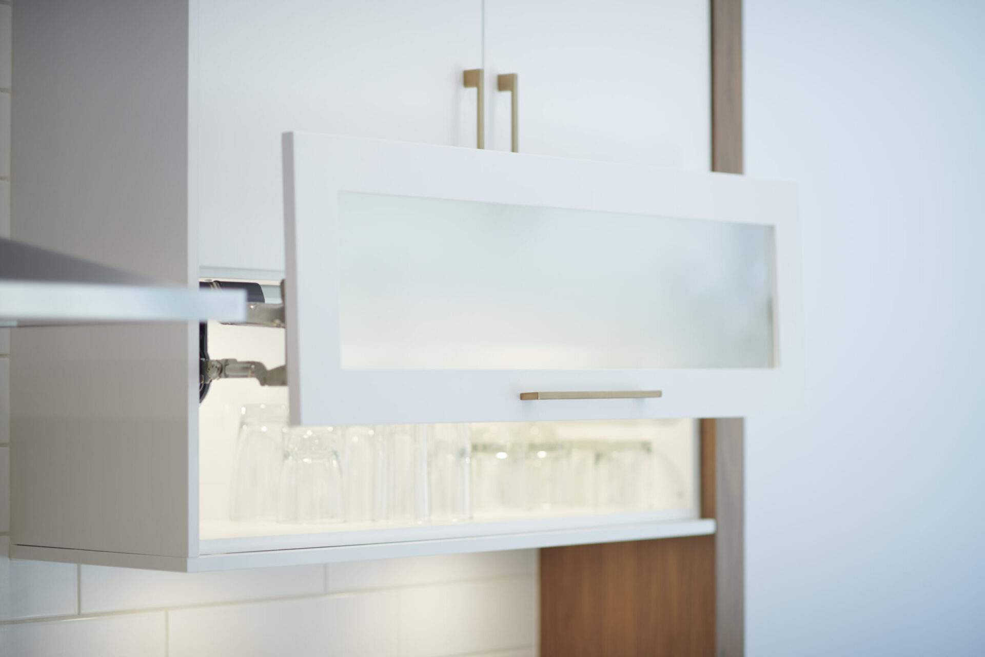 This image shows a modern, minimalist kitchen cabinet with a frosted glass front, partially open to reveal neatly arranged clear drinking glasses.