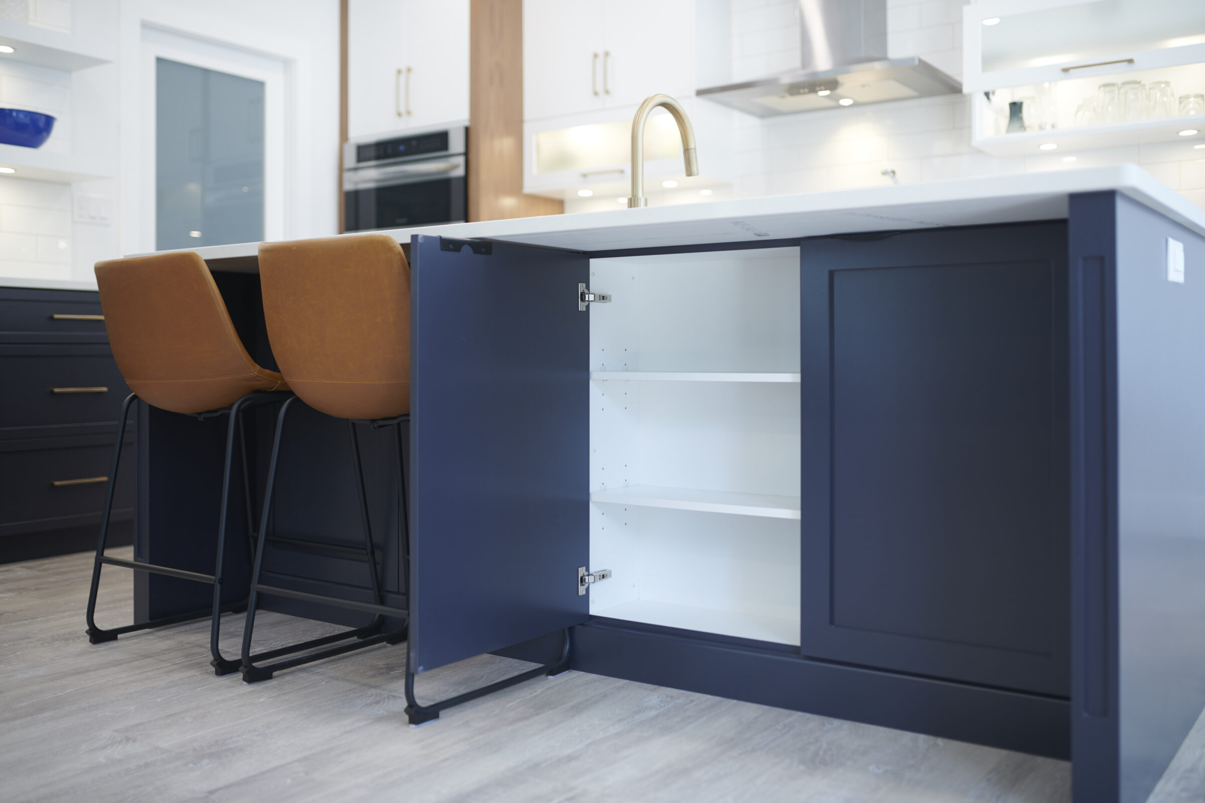 A modern kitchen with white countertops, navy blue cabinets, and two amber bar stools. An open cabinet door reveals empty shelves inside.