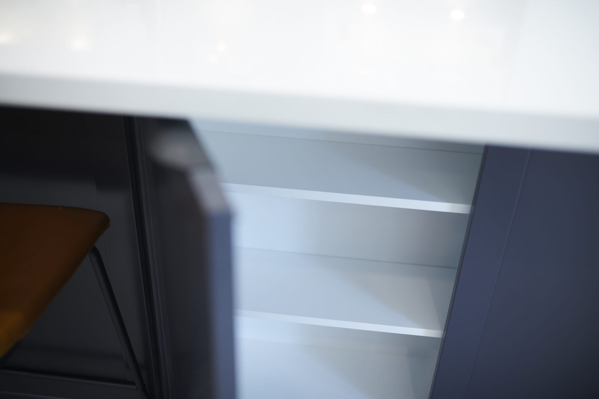 This image shows a modern, minimalist interior with an empty white shelf, dark cabinets, and a glimpse of a chair with a brown seat.