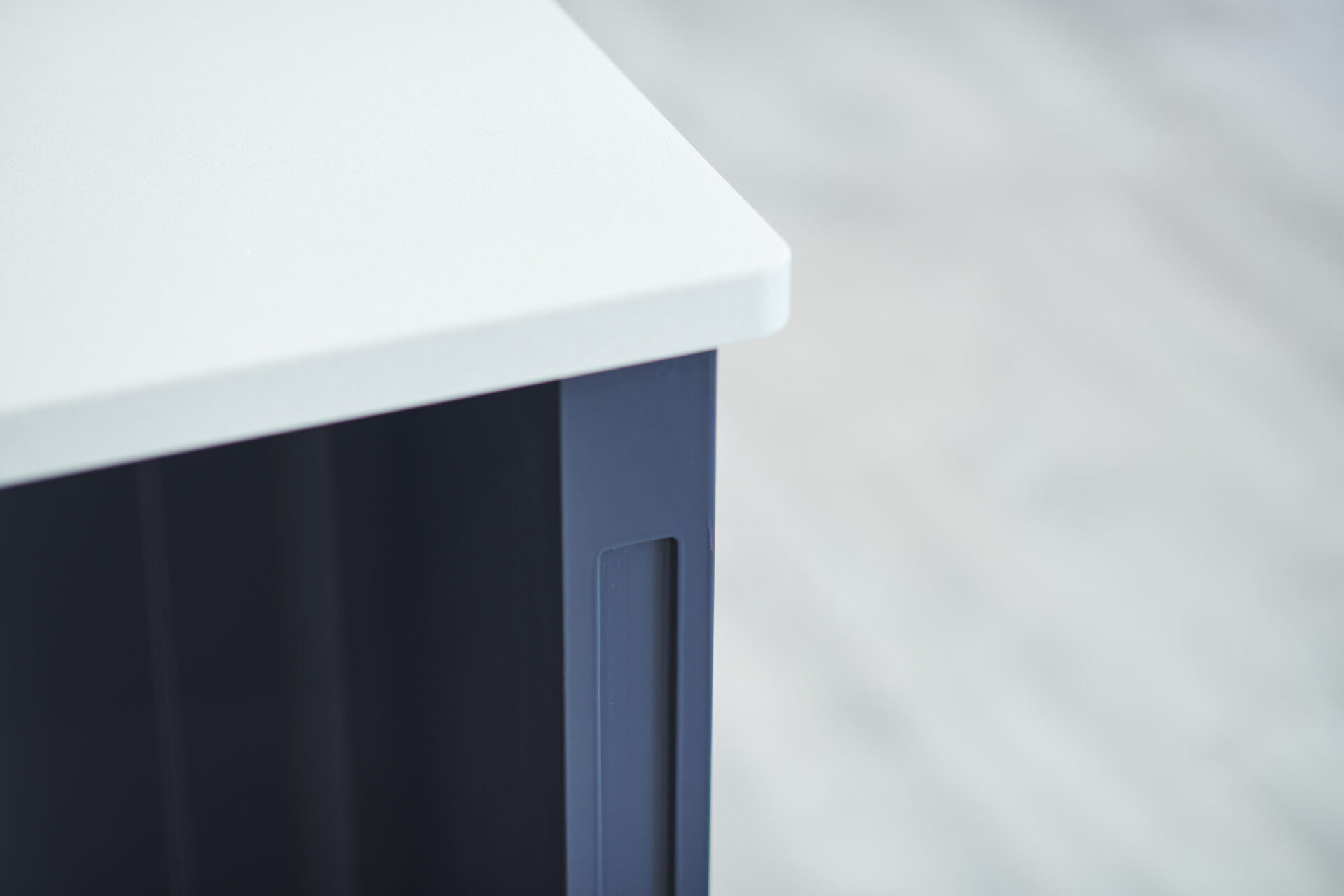 A corner of a modern desk with a white top and navy blue side panel featuring a sleek handle, set against a blurred light background.