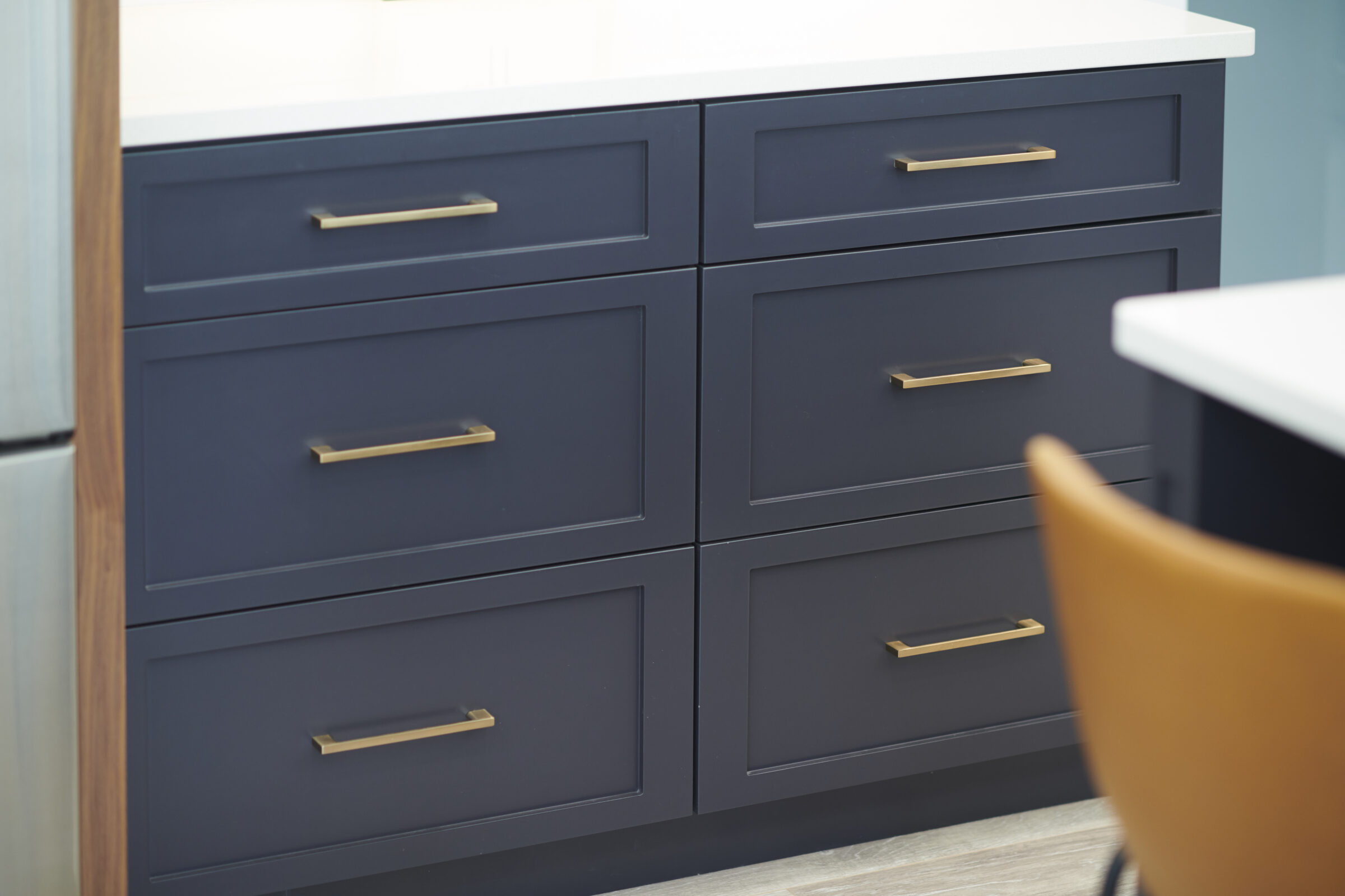 A modern blue cabinet with six drawers and brass handles sits next to a partial view of a white table and a tan chair.