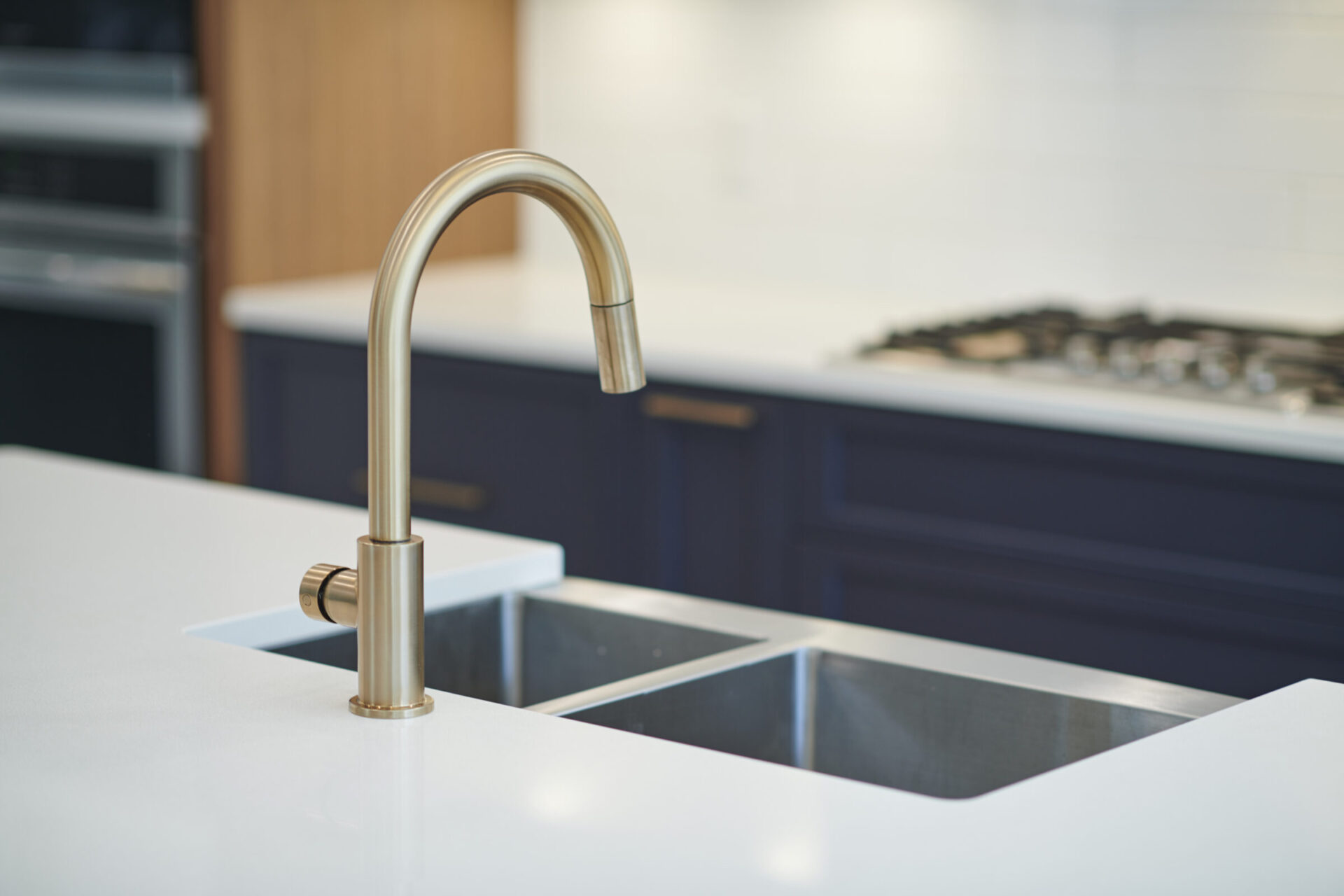 A modern kitchen with a brass faucet over a stainless steel sink, white countertops, and dark blue cabinets blurred in the background.