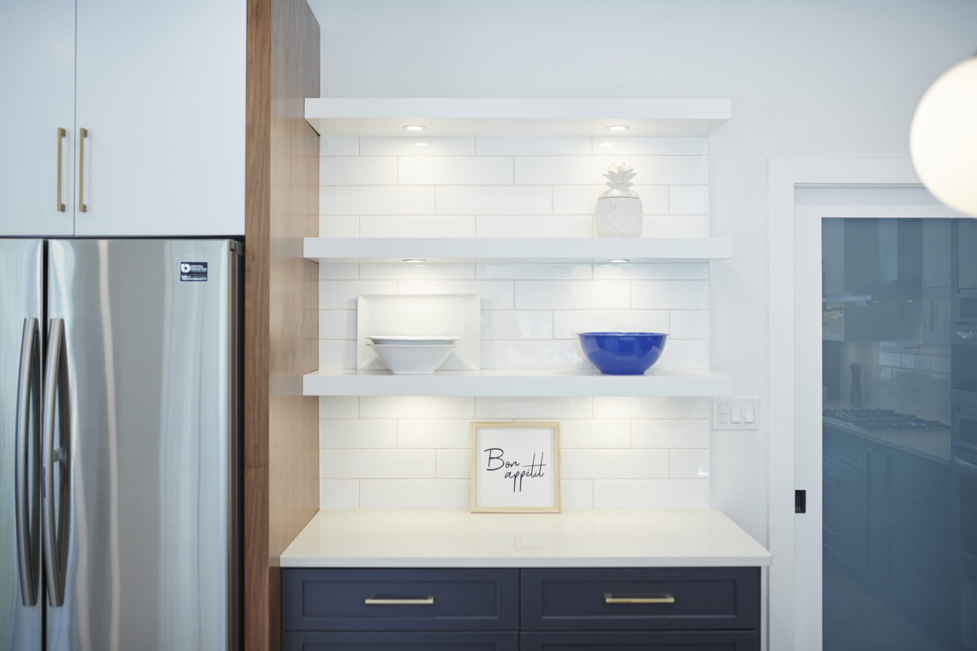 A modern kitchen corner features white subway tiles, floating shelves with minimal decor, a stainless steel refrigerator, and dark blue cabinets.