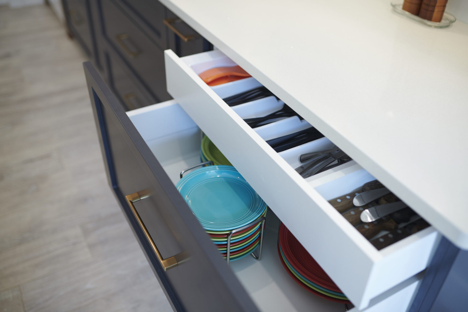 A modern kitchen drawer is open, showing neatly arranged colorful plates, bowls, and assorted cutlery, set against dark cabinets and a light countertop.