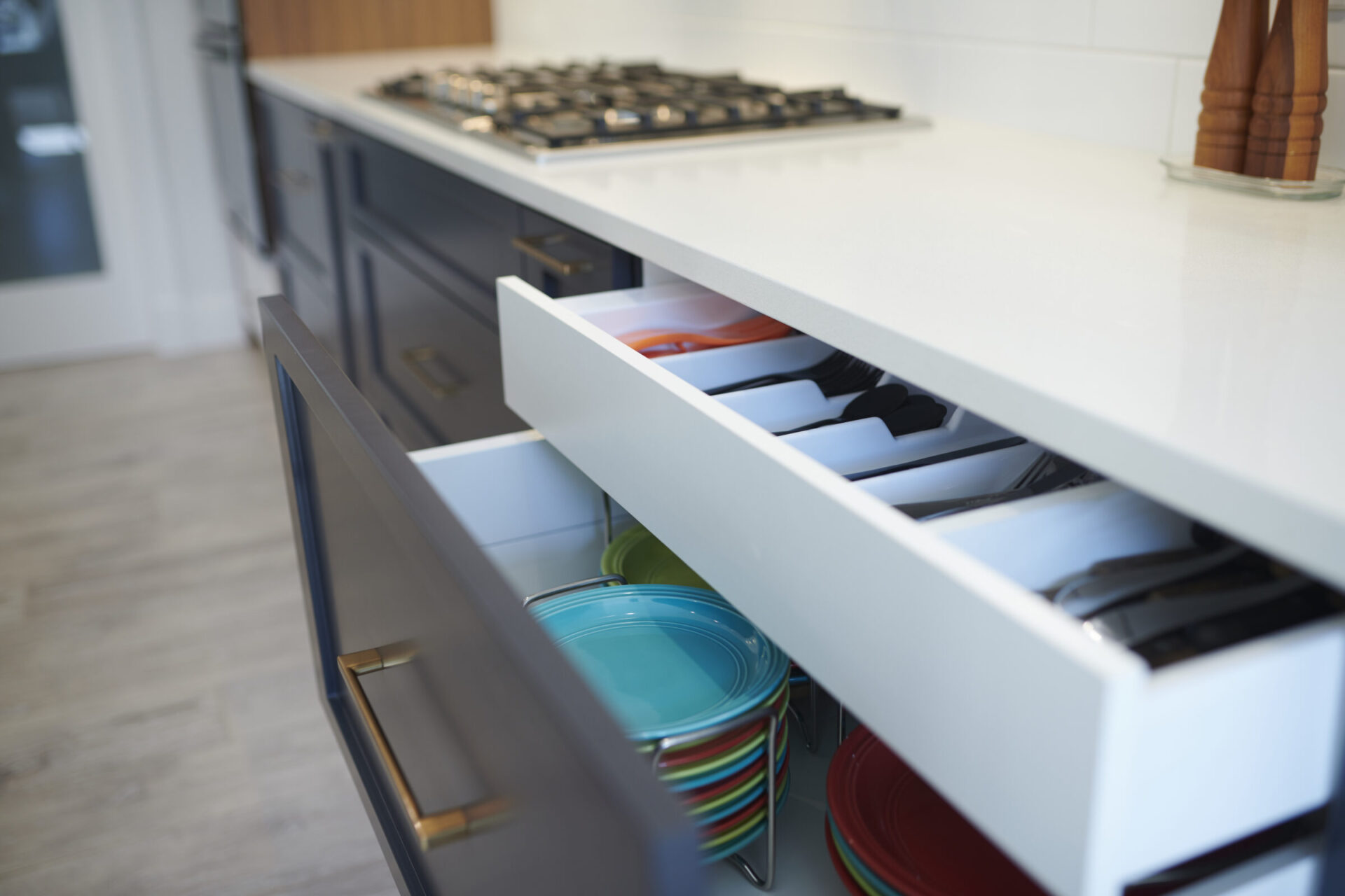 Modern kitchen interior with an open drawer revealing organized plates and utensils. Gas stove and neutral color scheme with a wooden floor visible.