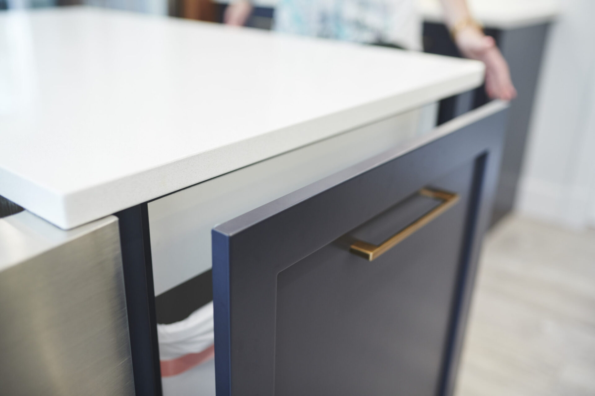 This image showcases a modern kitchen counter with a sleek white top, navy cabinets, gold handles, and a partial view of a person nearby.