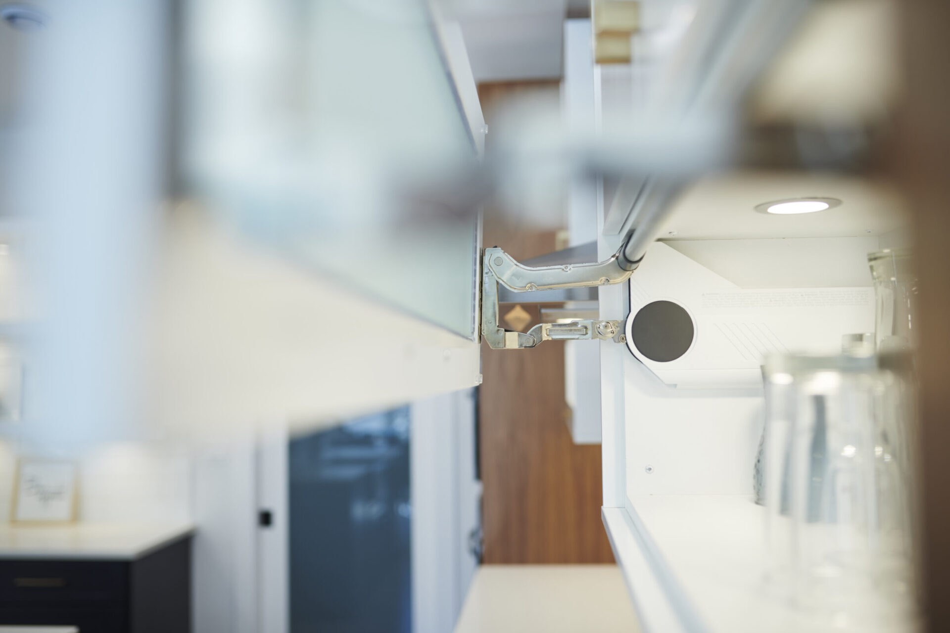 This image shows an interior view focusing on an open cabinet hinge in a modern kitchen, with blurred glasses in the foreground and cabinetry in the background.