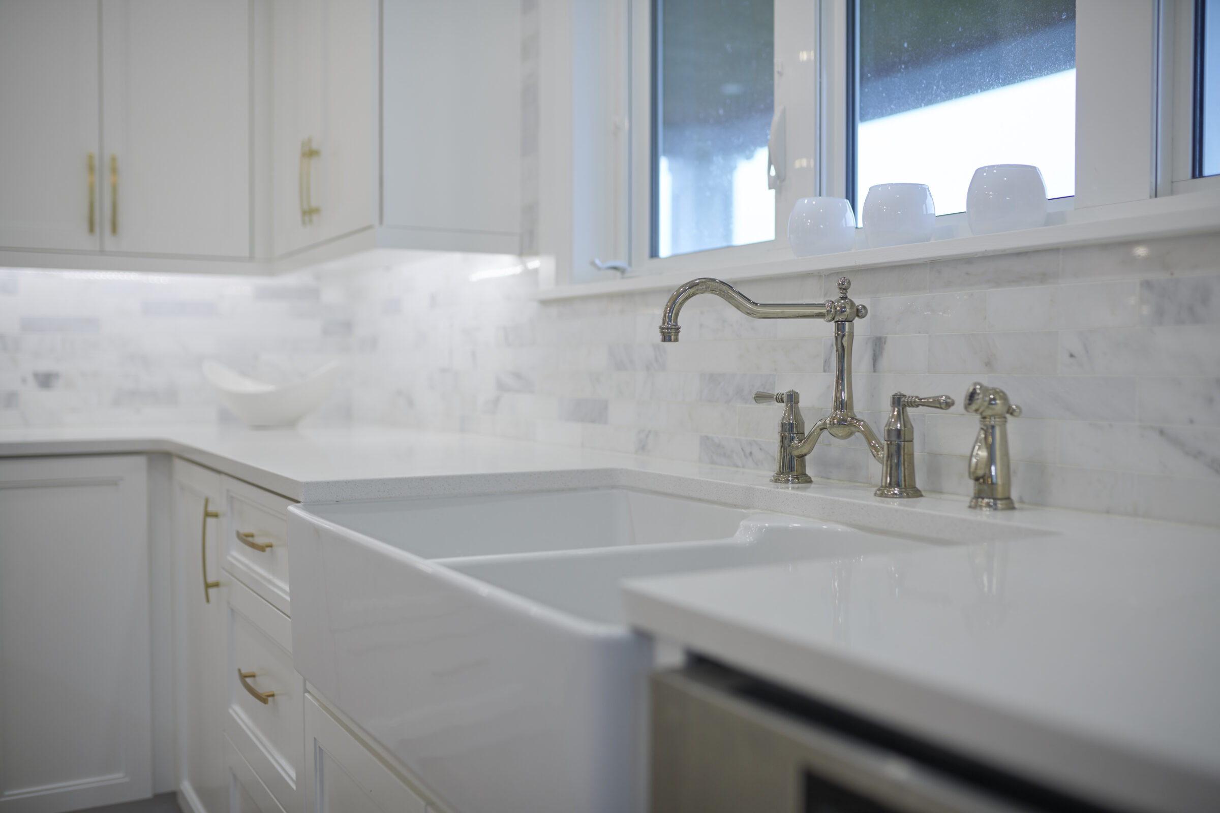 A modern kitchen featuring white cabinetry, marble backsplash, a farmhouse sink, and sleek gold handles, with a view of the window.