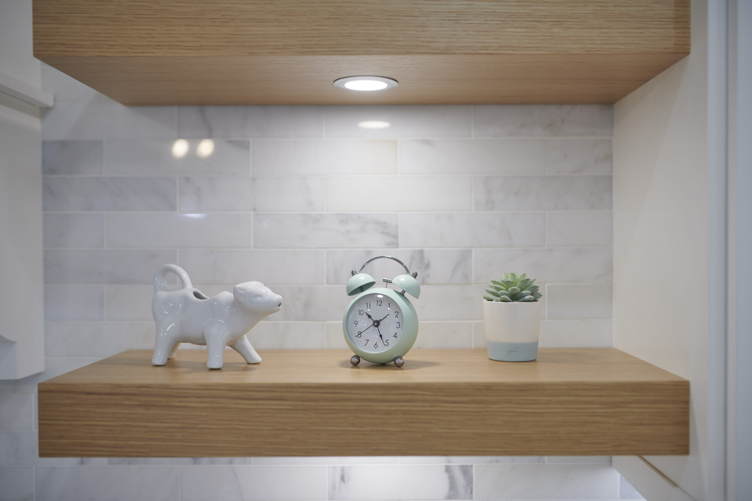 A minimalist shelf with a porcelain cat figurine, an aqua-colored alarm clock, and a potted succulent plant. White marble tiles and wooden accents.