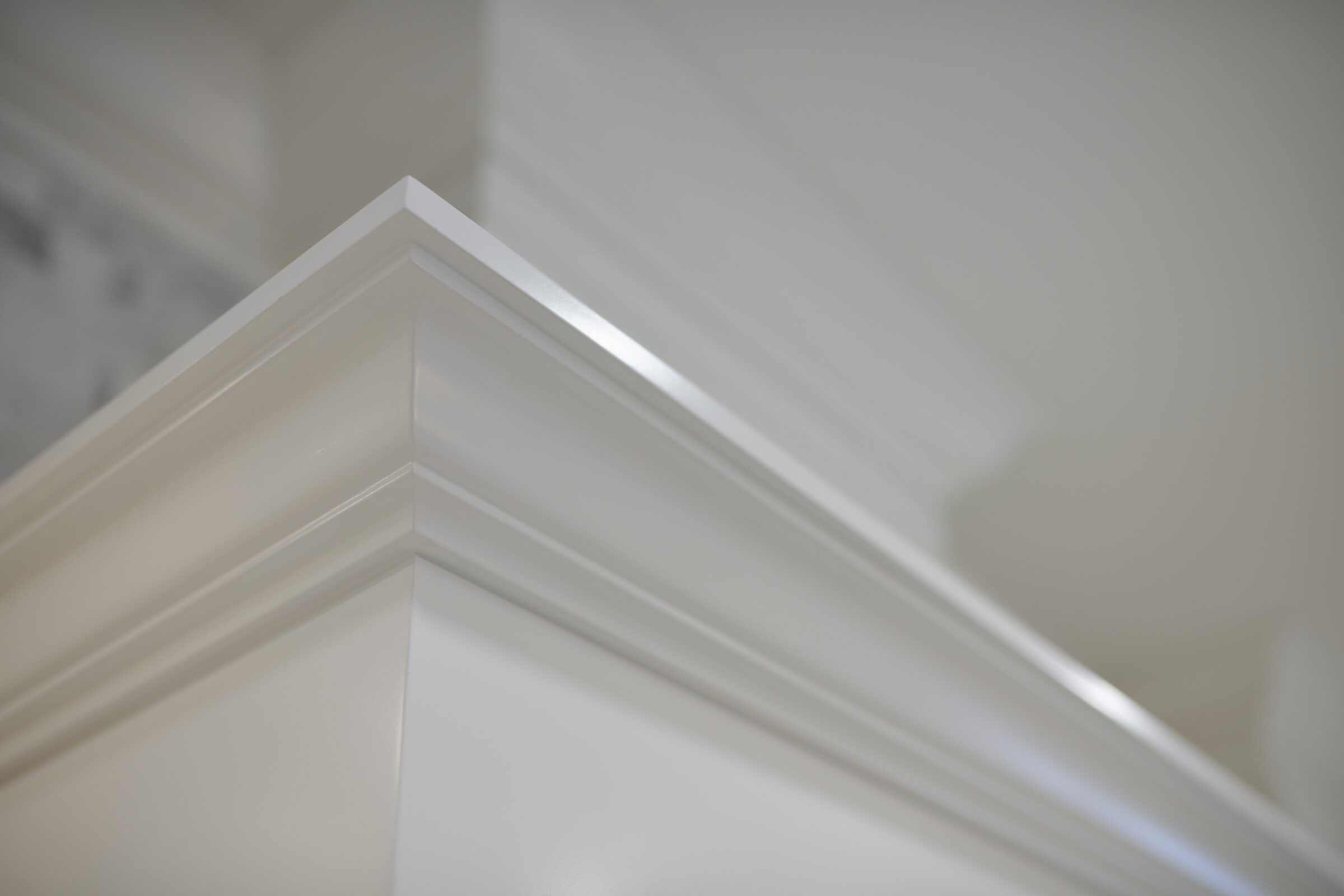 This image shows a close-up of white crown molding at the junction where a ceiling meets a wall, with a soft-focus background and subtle shadowing.