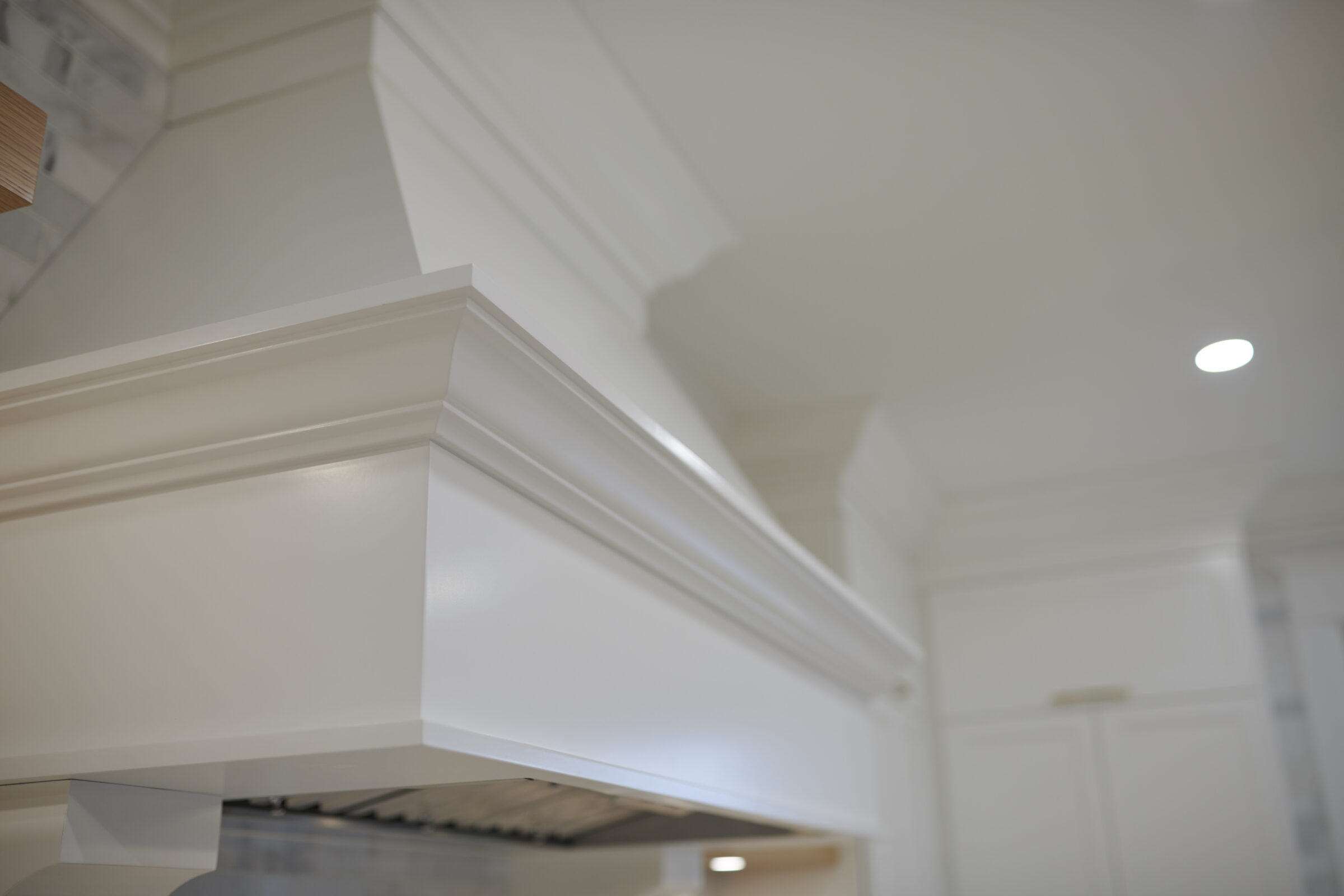 The image shows a white interior with detailed crown molding design. A recessed light fixture illuminates the neutral-toned ceiling space, hinting at a modern kitchen.