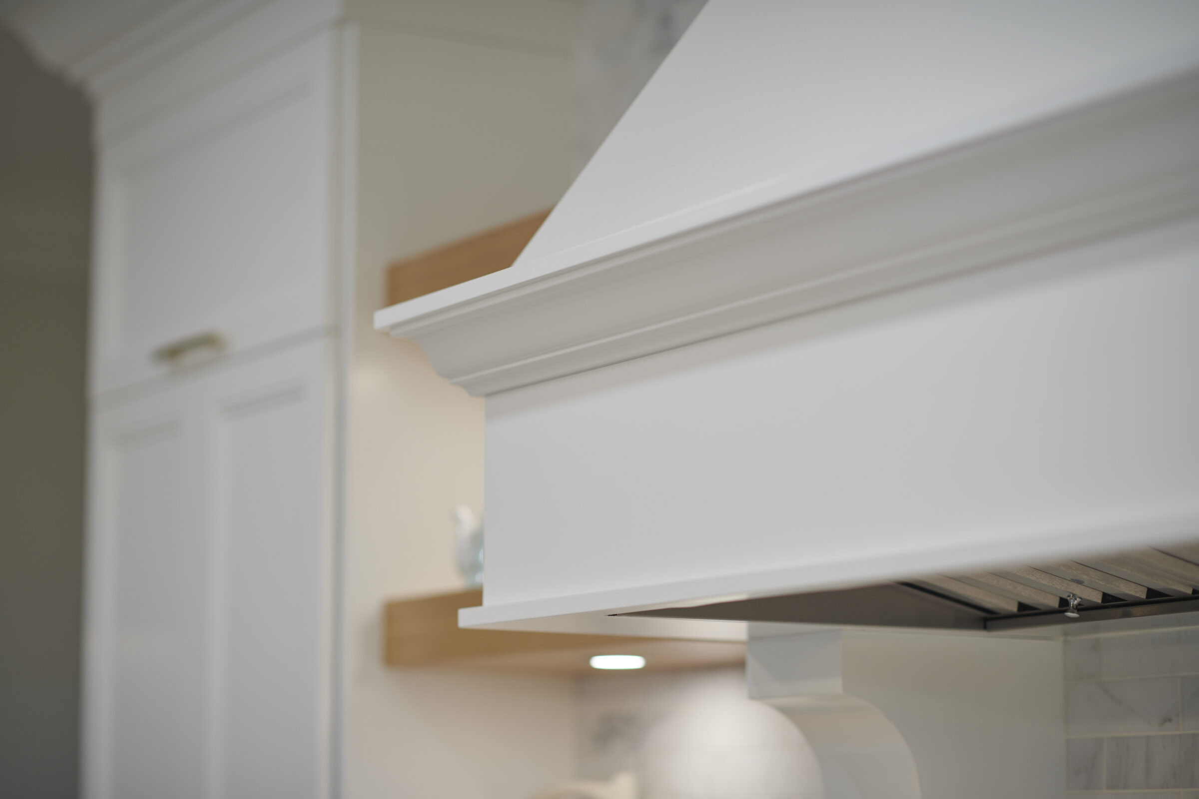 A close-up photo of a white kitchen hood with a glimpse of the wooden cupboard above and a cabinet in the soft-focus background.