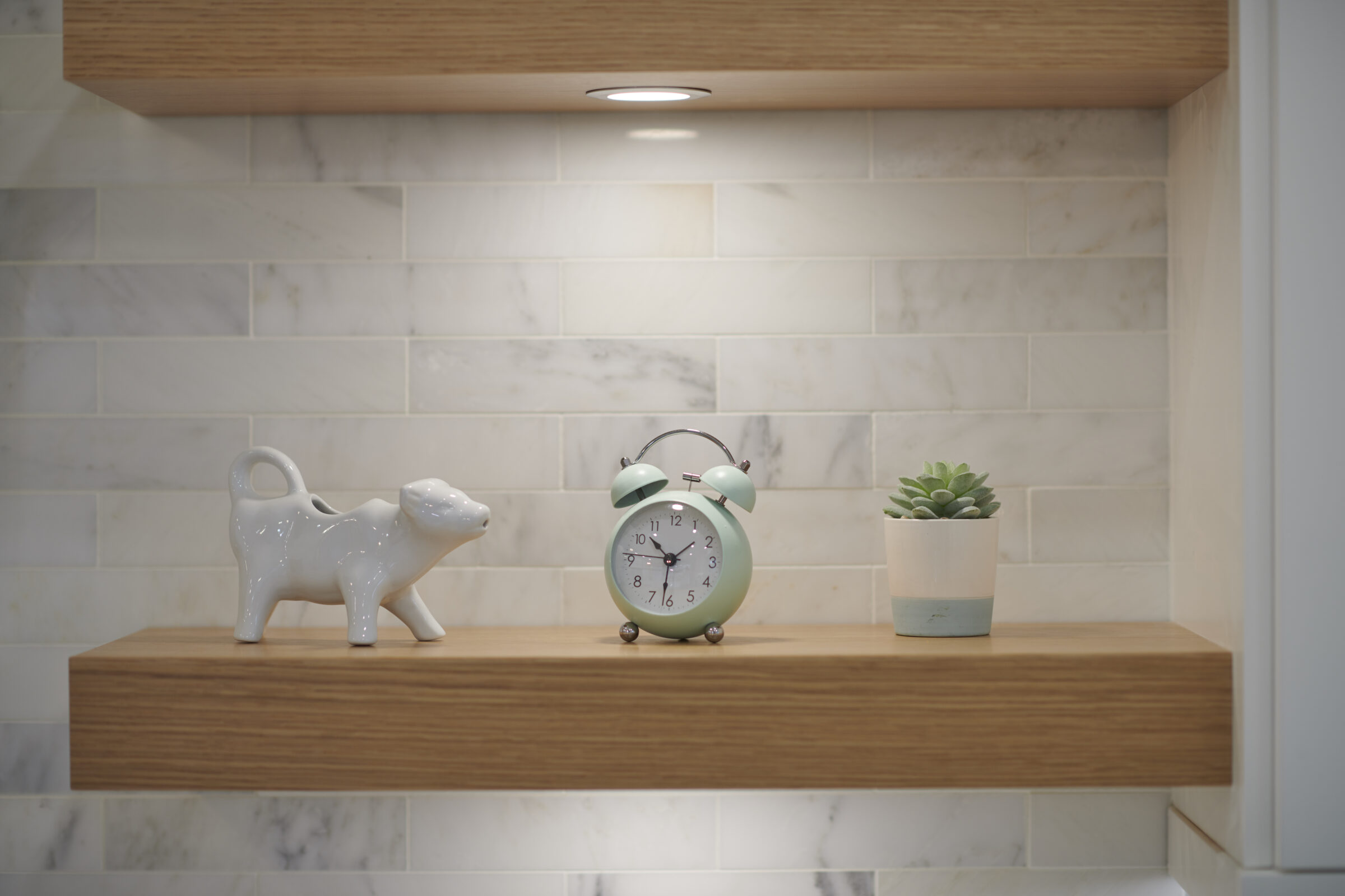A wooden shelf against a marble tiled wall displaying a ceramic sheep figurine, a retro green alarm clock, and a potted succulent plant.