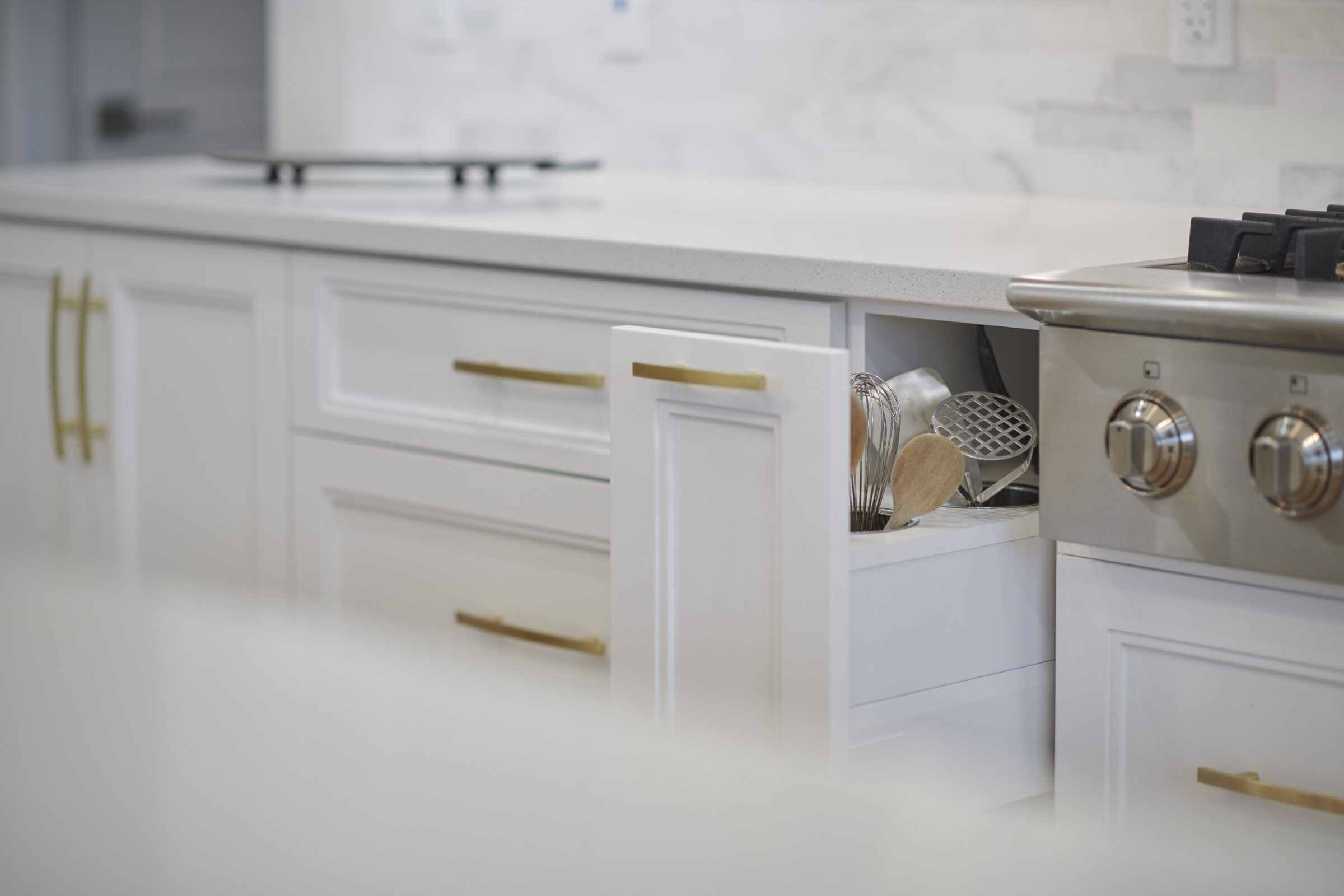 A modern kitchen with white cabinets, gold handles, a cooktop, and cooking utensils peeking out from an open drawer. It has a bright and clean aesthetic.