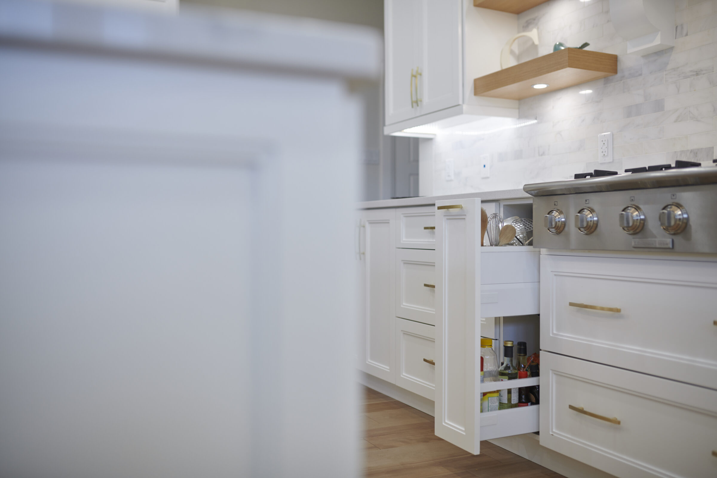 A modern kitchen with white cabinetry, stainless steel stove, open drawer revealing bottles and utensils, and a marble backsplash with wooden shelves.