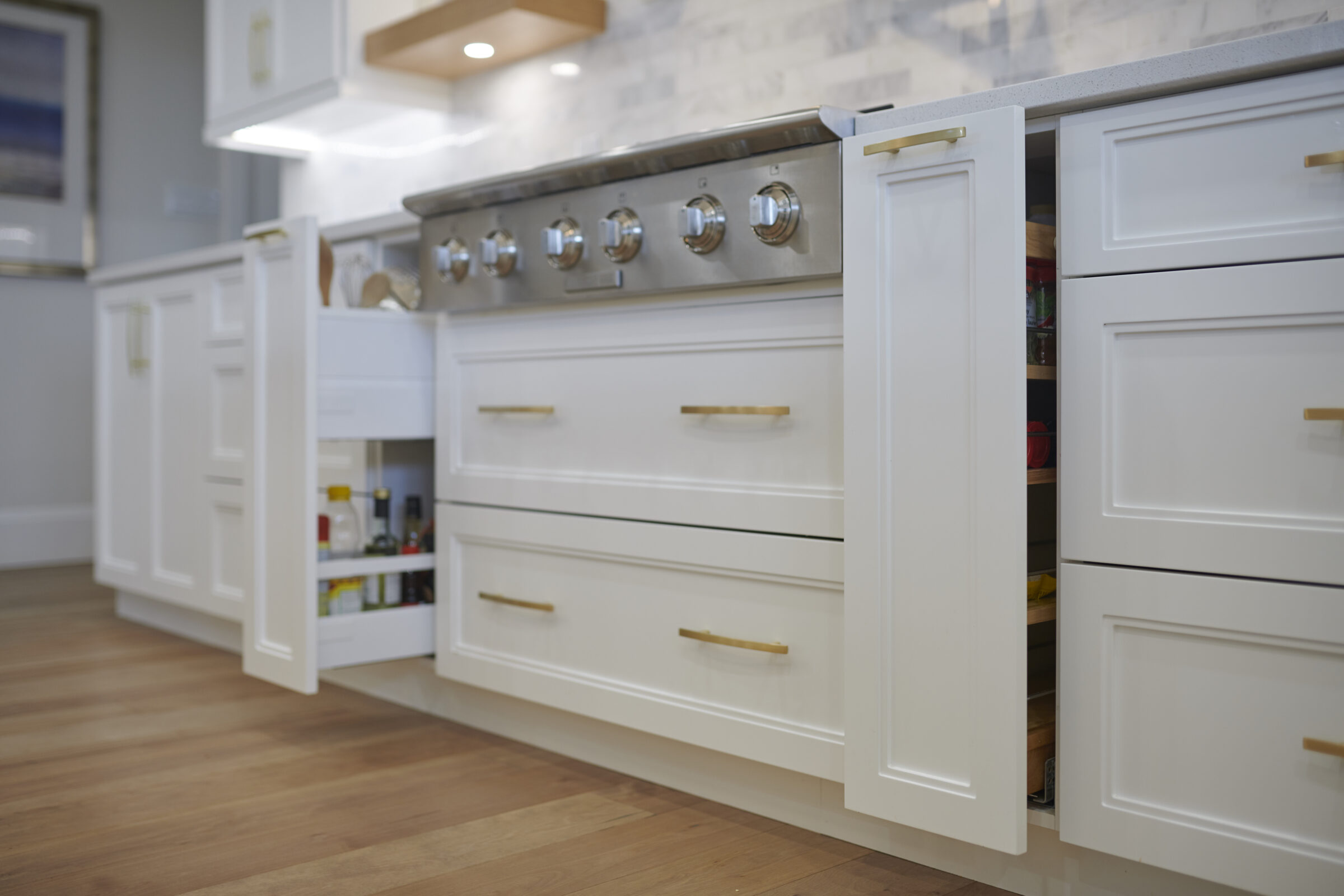 Modern kitchen with white cabinetry, stainless steel stove, gold handles, and hardwood floor. One cabinet is open, revealing contents inside.