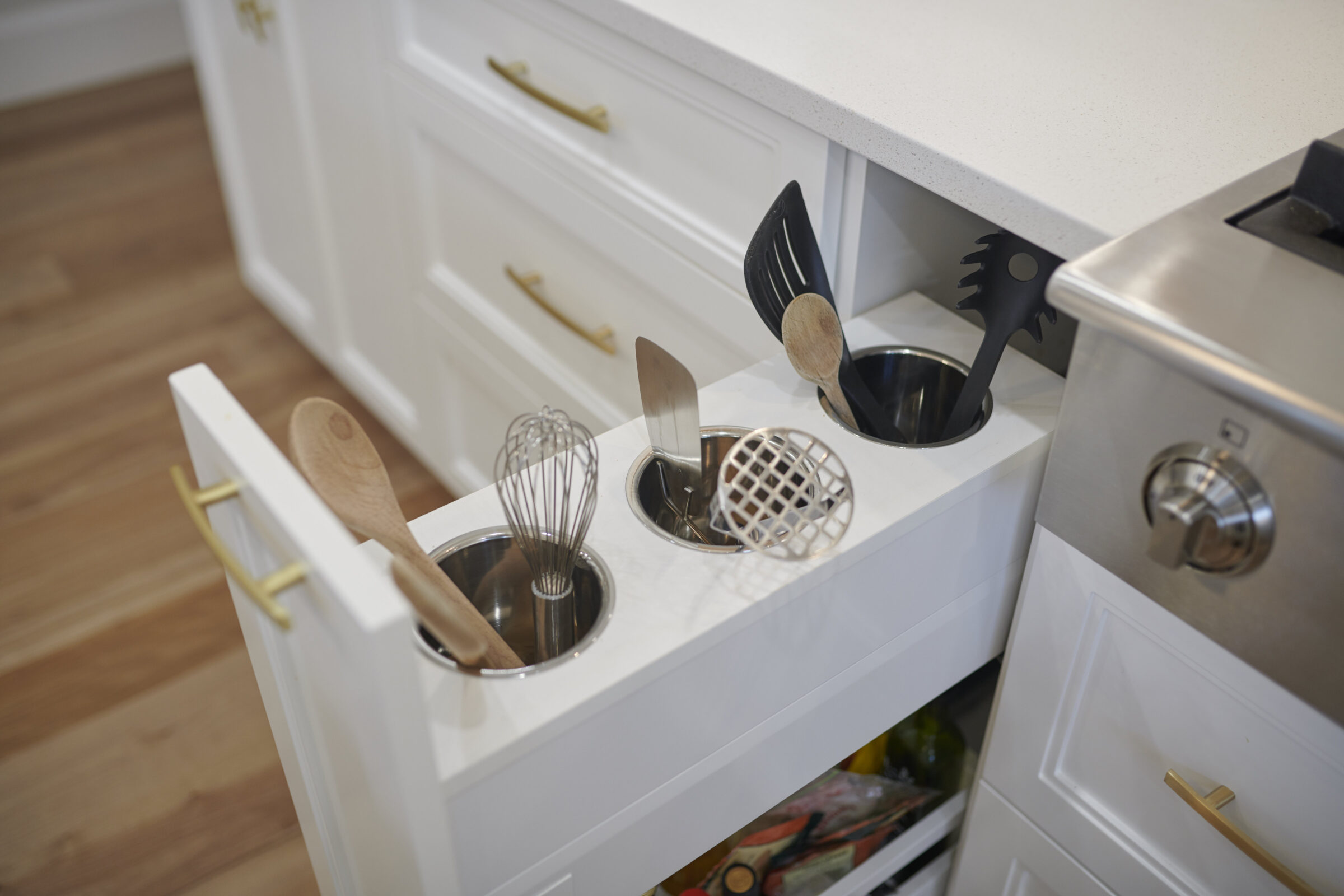 A modern kitchen with white cabinetry featuring gold handles. An open drawer contains various cooking utensils neatly organized in stainless steel cups.
