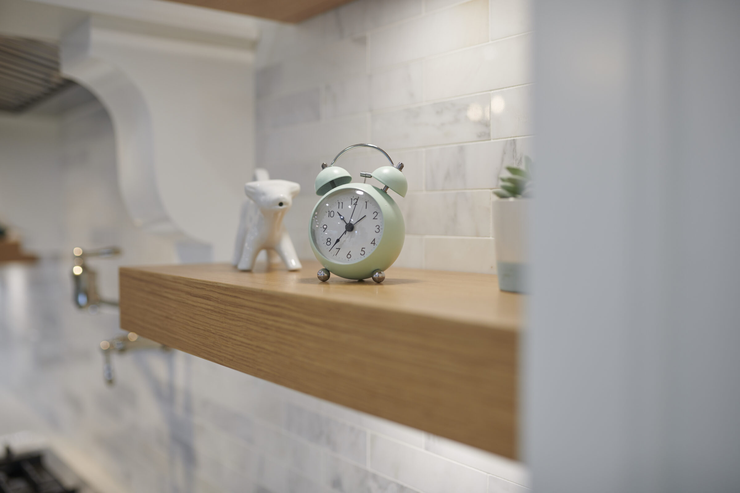A mint green analog alarm clock sits on a wooden shelf, alongside a small, white figurine and a potted plant, against subway tile backsplash.