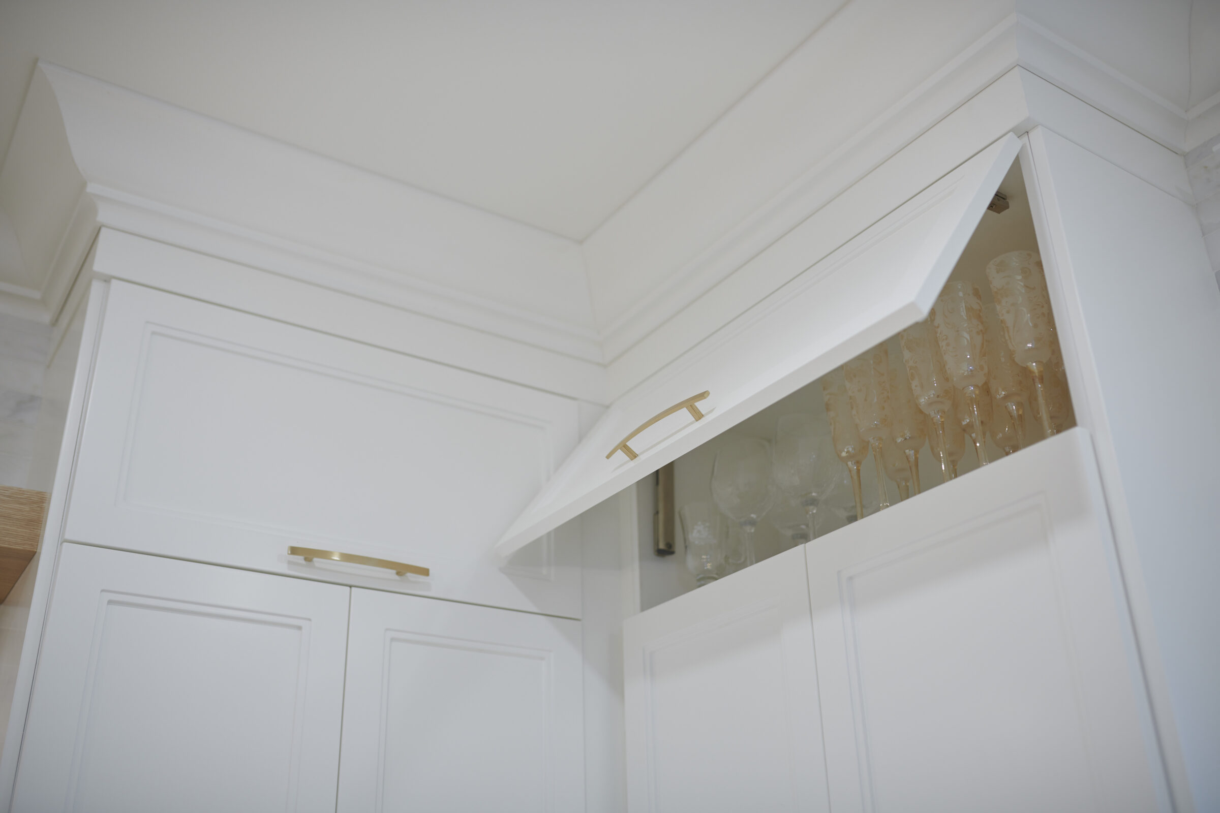 The image shows a modern white kitchen cabinet with a tray ceiling and glassware visible inside an overhead compartment with gold handles.