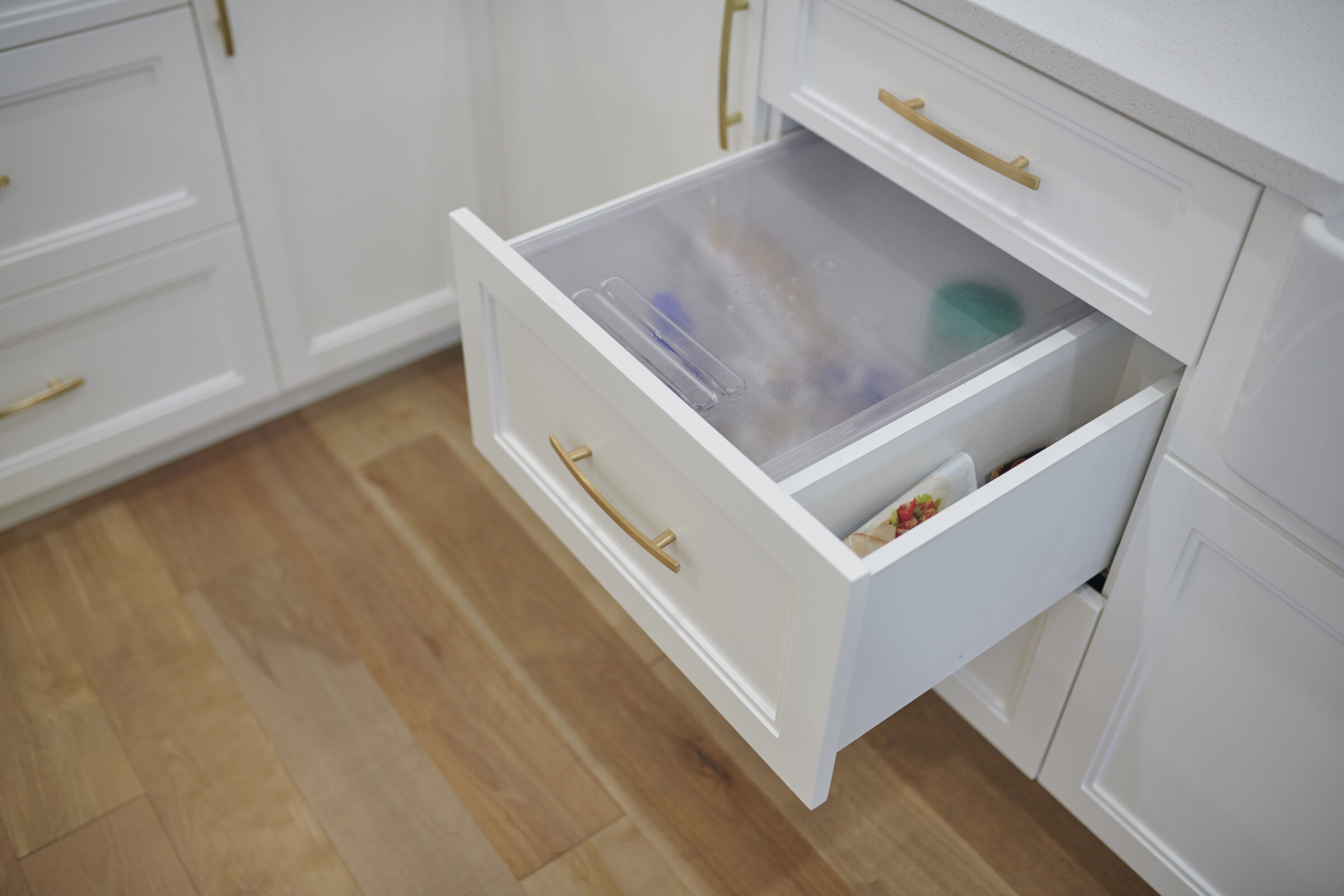 An open white kitchen drawer with gold handles reveals a frosted organizing tray with utensils and compartments. The modern cabinetry contrasts with wooden flooring.