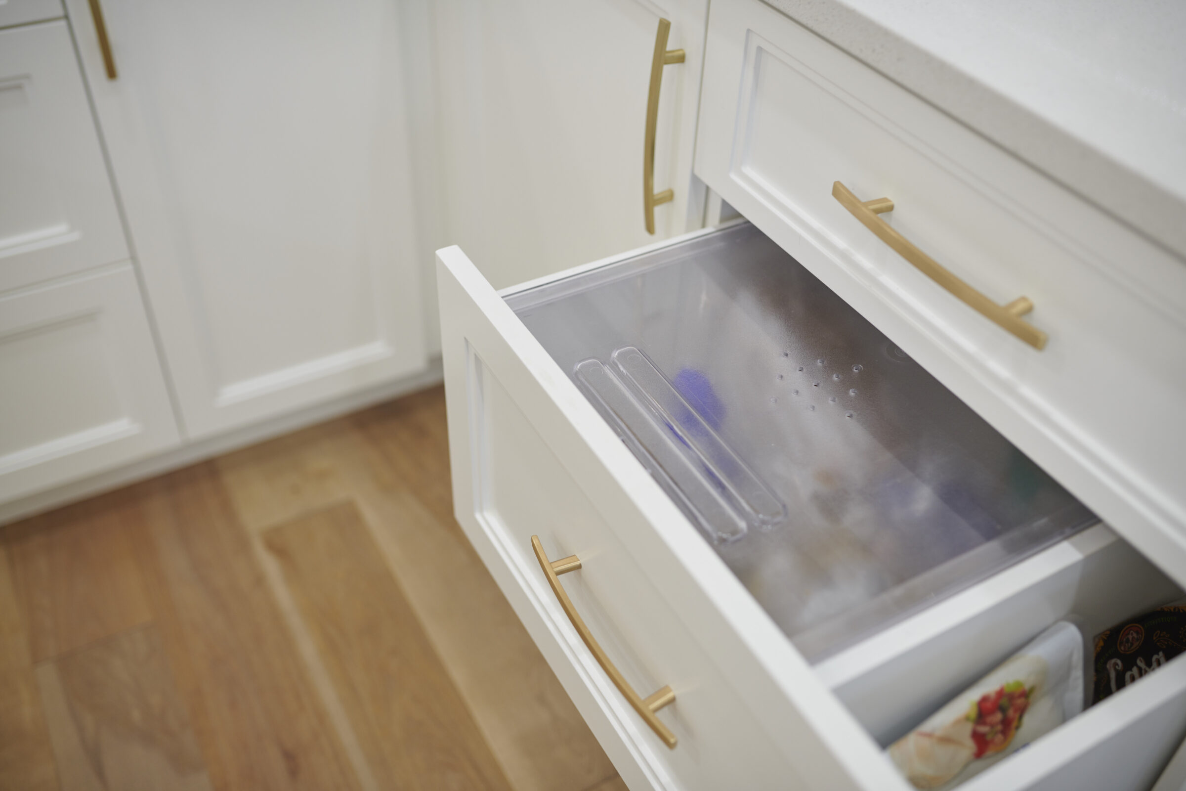 A modern kitchen interior with a white freezer drawer open, revealing a frosted glass shelf and frozen food items inside. Wooden floor and gold handles visible.