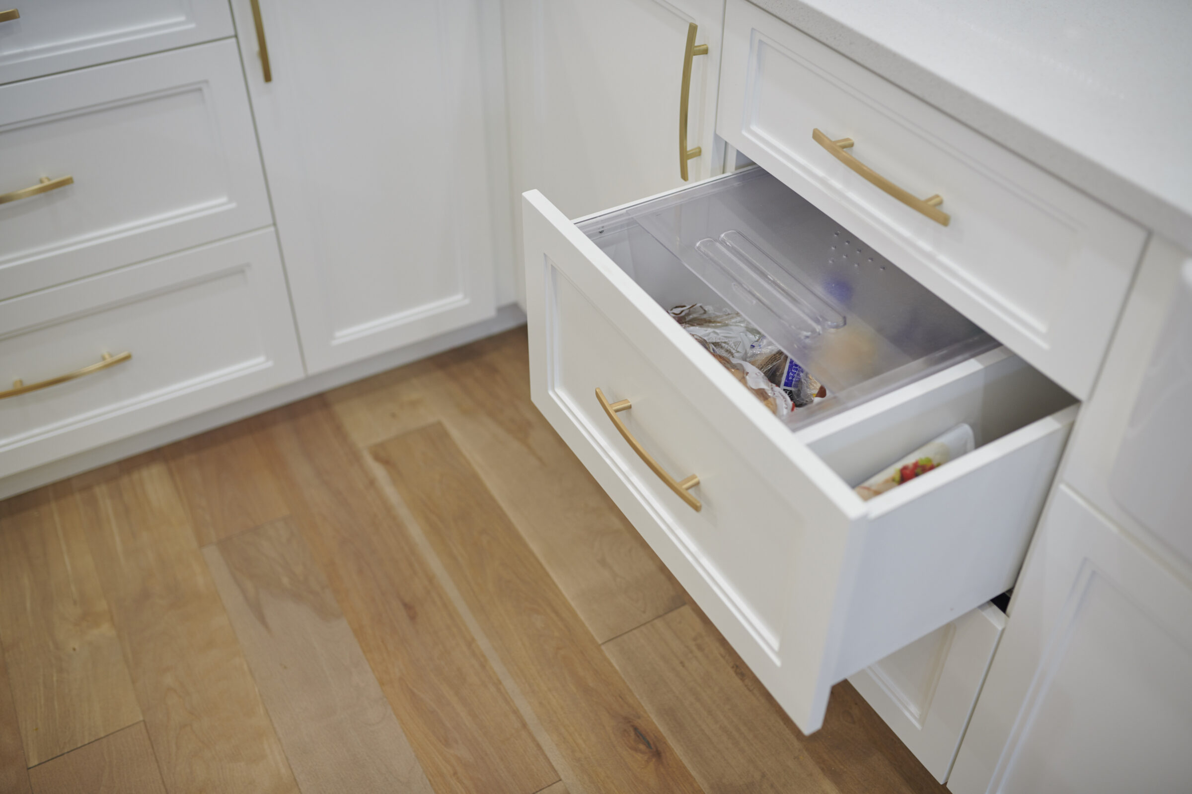 A modern kitchen with white cabinets featuring gold handles, a wooden floor, and an open freezer drawer containing frozen foods and ice packs.