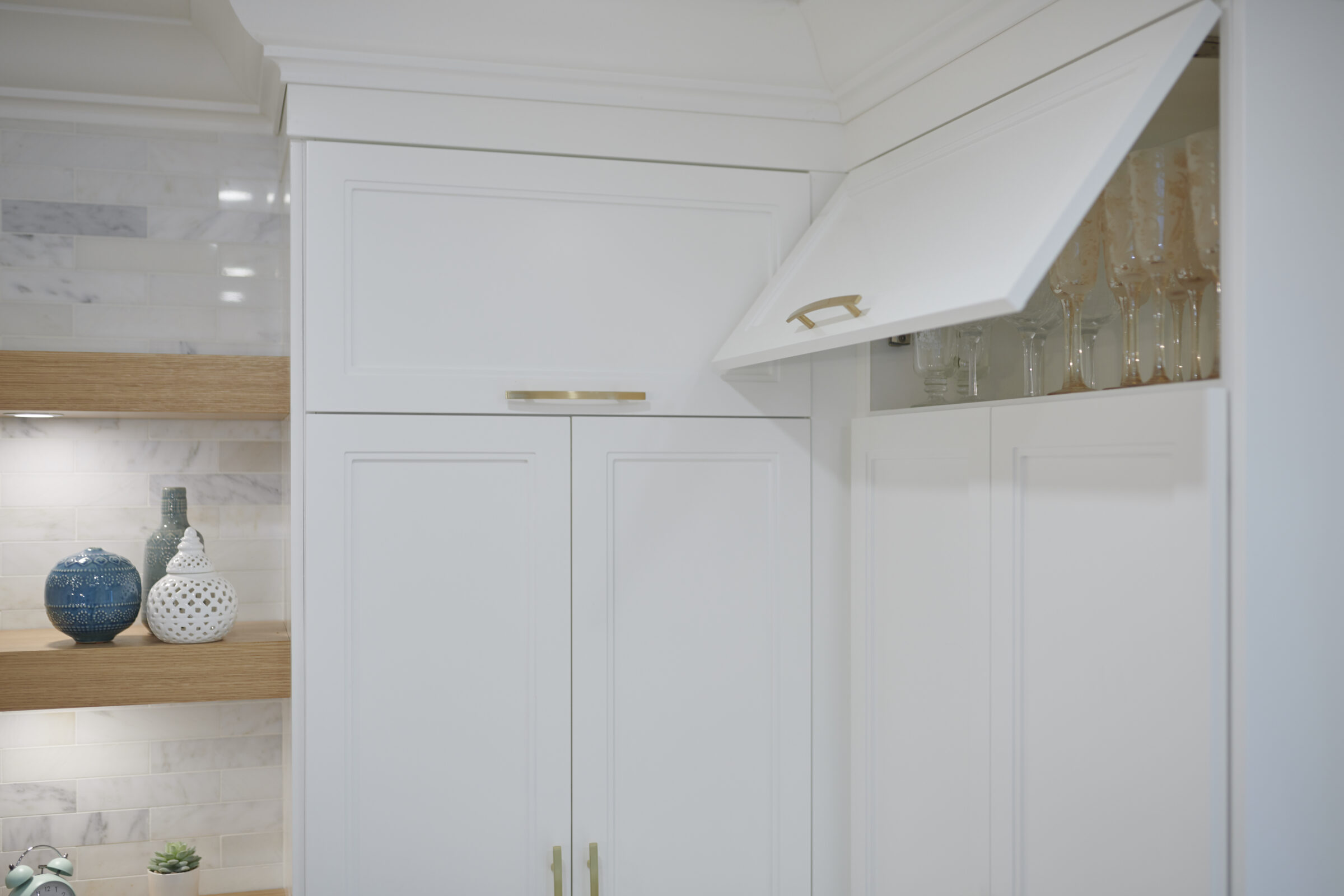 A modern kitchen with white cabinetry, gold handles, marble backsplash, and wooden shelves displaying decorative vases. Wine glasses hang neatly above the counter.