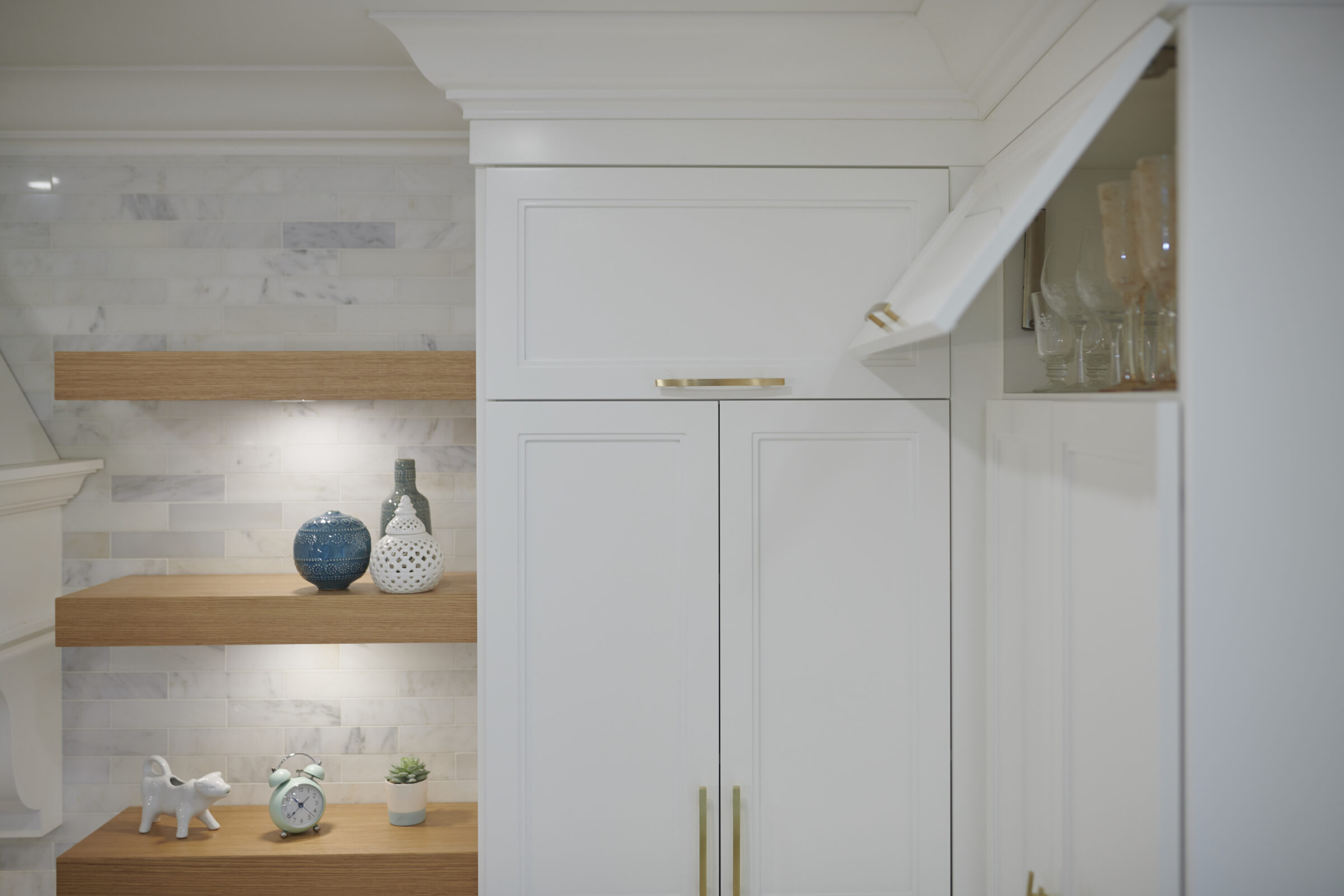 The image shows a modern kitchen corner with white cabinetry, marble backsplash, wooden shelves adorned with decorative items, and gold-colored hardware.